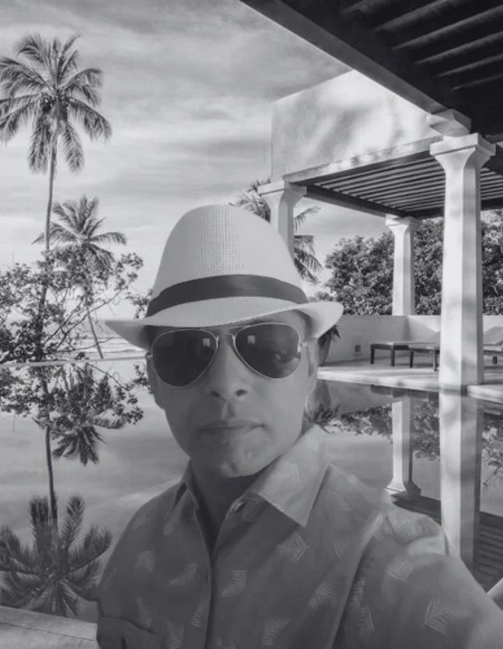 Man wearing a fedora hat, sunglasses, and a patterned collared shirt taking a selfie outdoors near a pool with palm trees and structures in the background.