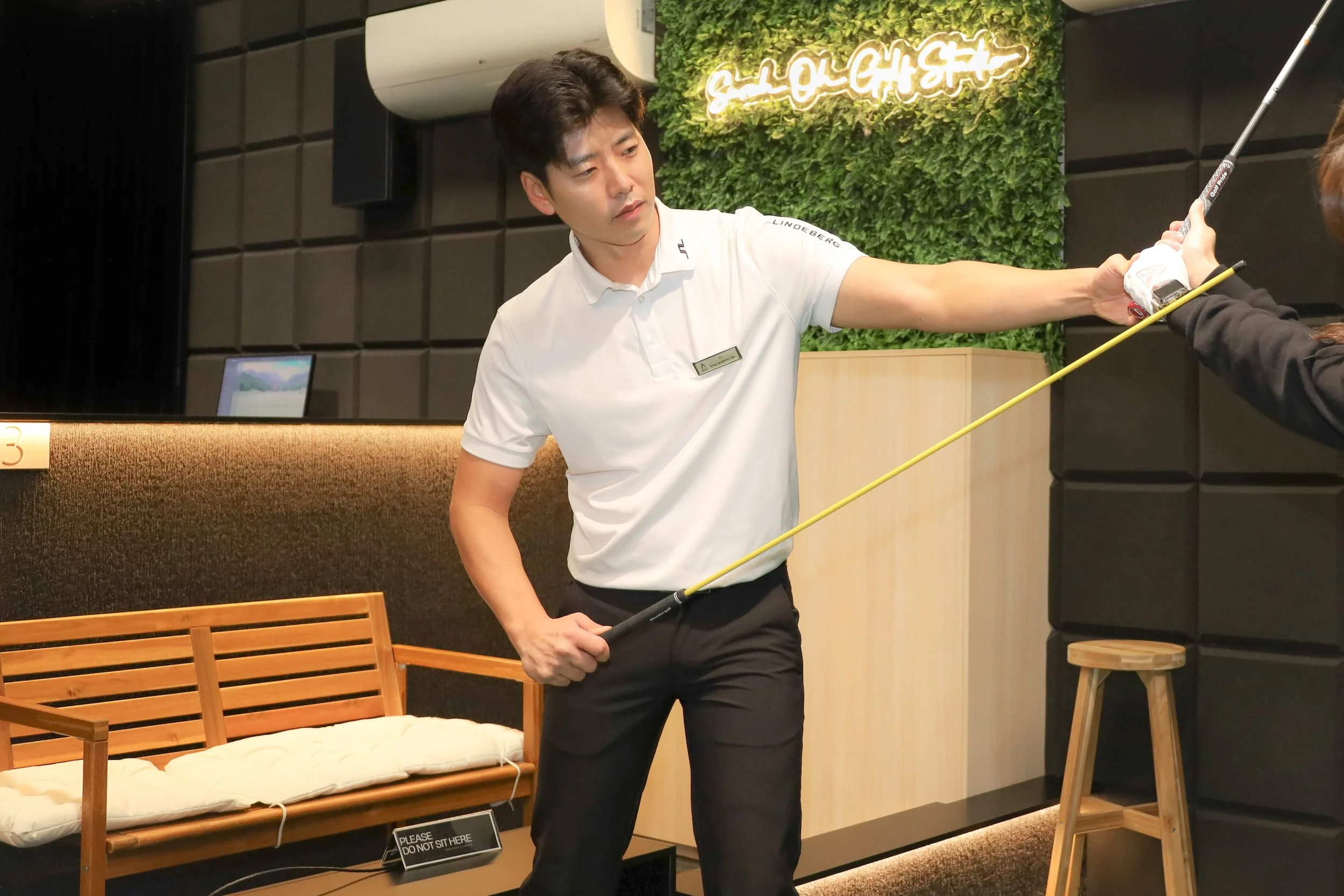 A man in a white polo shirt with a name tag is practicing golf indoors, aiming a golf club with a yellow shaft at a screen located in a hospitality or lounge area with black walls and green decorative wall. There is a bench with a sign that says "Please do not sit here."