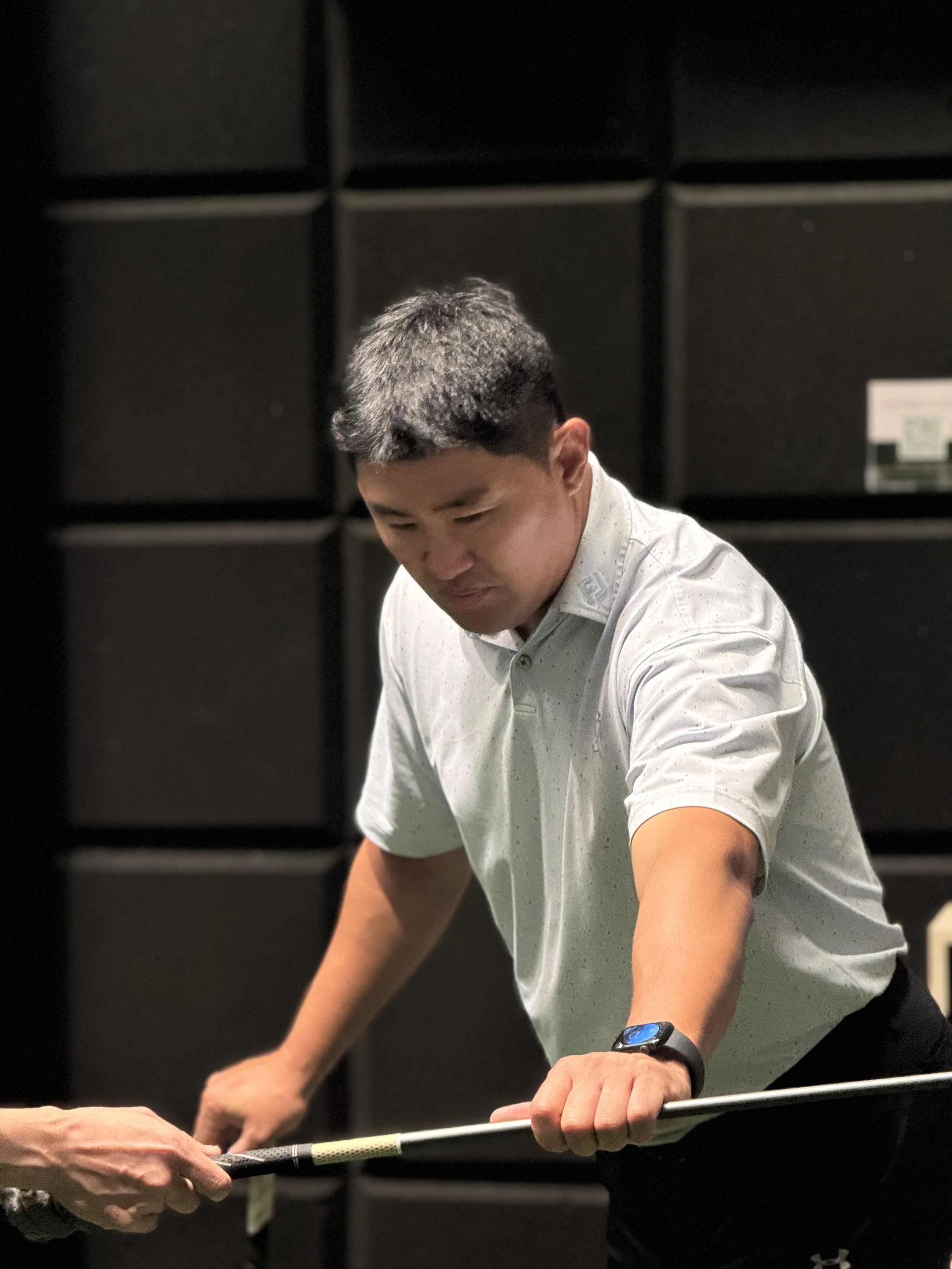 A man wearing a light gray polo shirt shows focus as he holds a golf club, in a room with black acoustic panels on the wall.