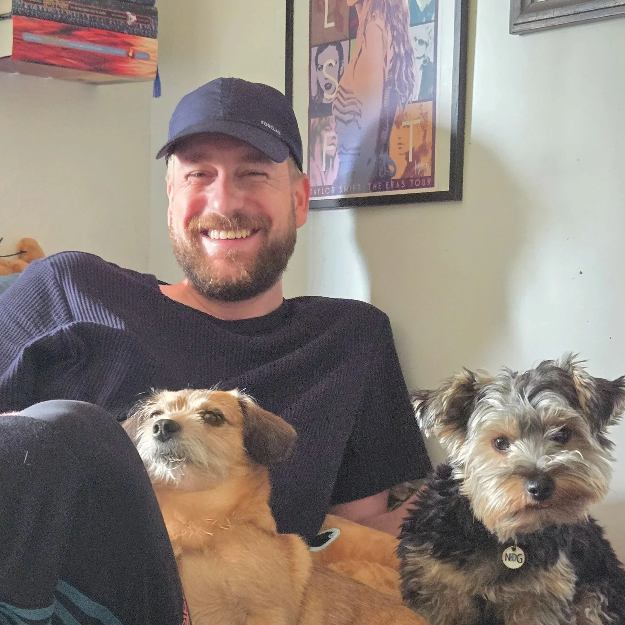 A smiling man with a beard holding a small brown and black puppy outdoors with trees and a small house in the background.