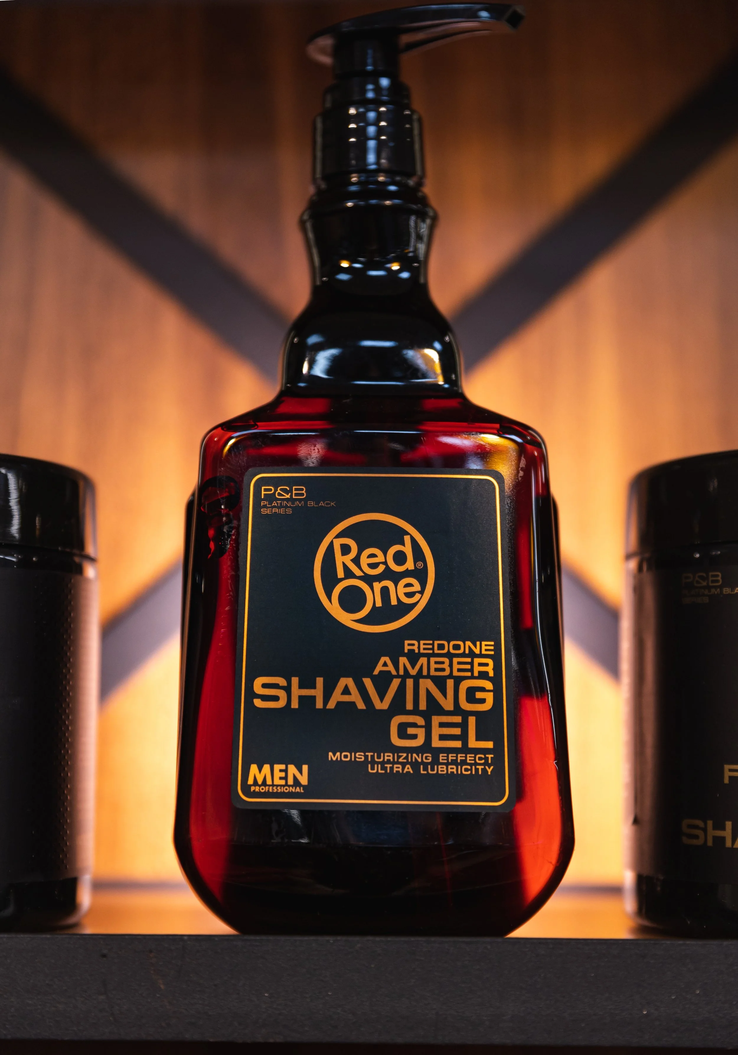 A bottle of Red One Amber Shaving Gel for men on display with a black pump top and a dark blue label with orange text, positioned between two other black containers.