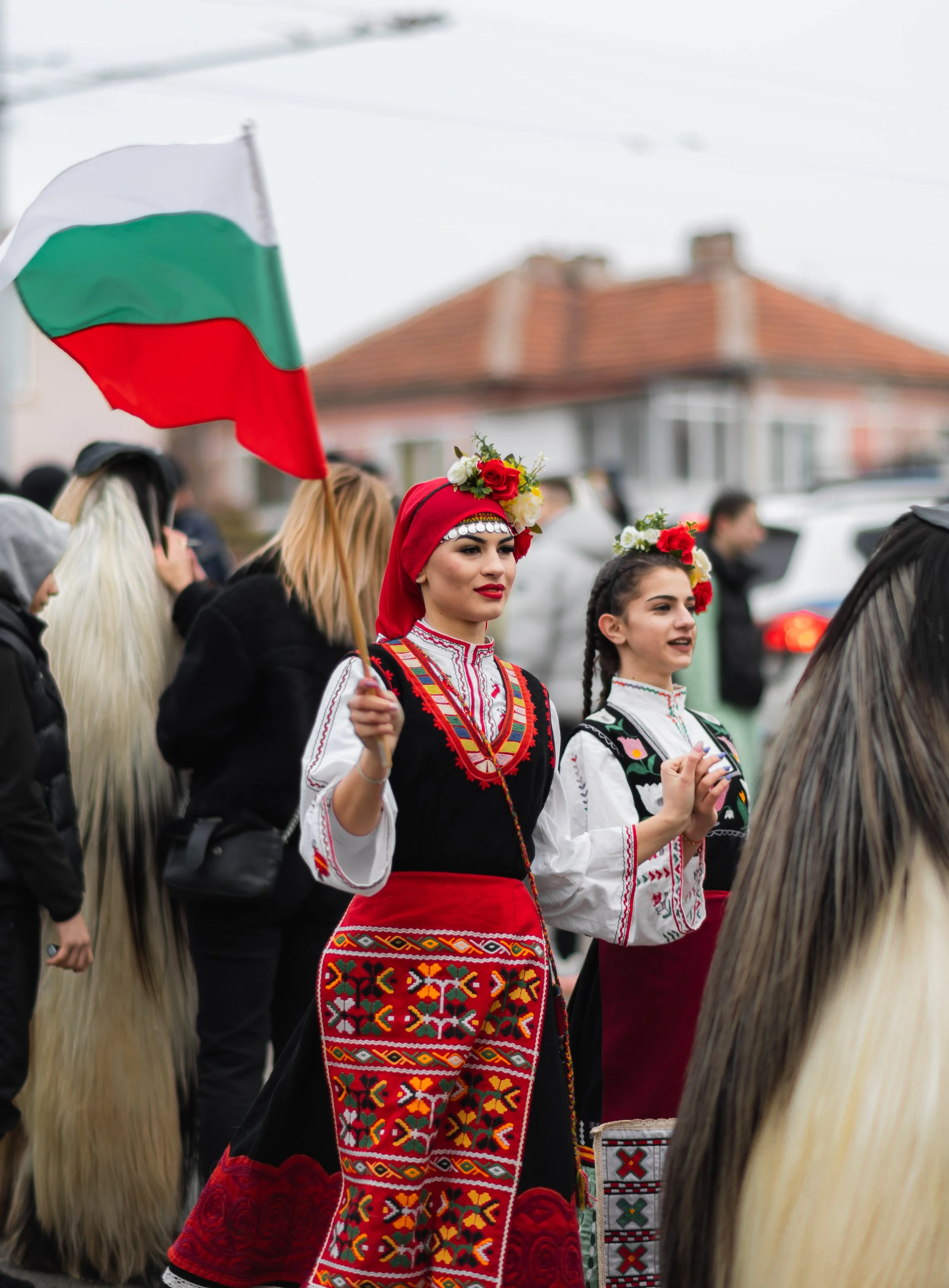 Two women dressed in traditional Bulgarian clothing, with floral headbands, participating in a parade, while holding the Bulgarian flag.