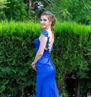 A young woman in a blue dress standing outdoors near a green hedge with trees in the background.