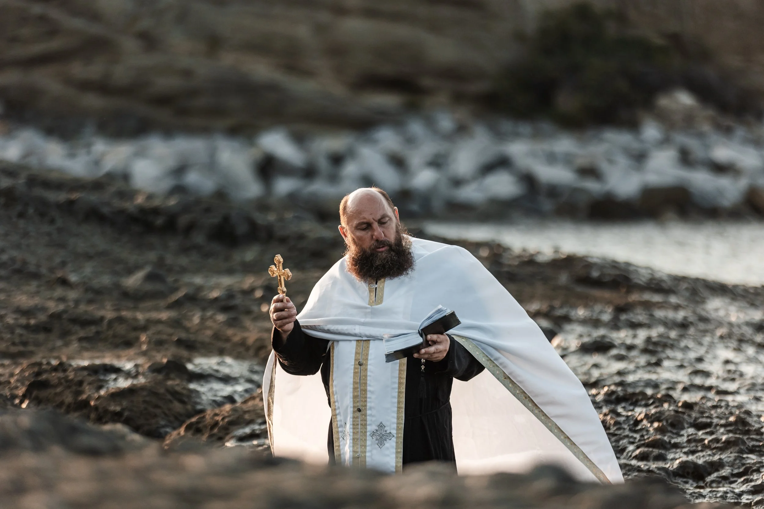 A man dressed as an Orthodox priest holding a cross and reading from a book outdoors near a rocky shoreline.