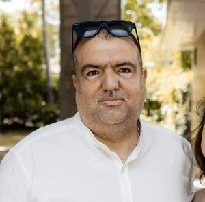 Close-up of a middle-aged man with short dark hair, wearing a white shirt, and sunglasses resting on his head, outdoors with blurred trees and a house in the background.