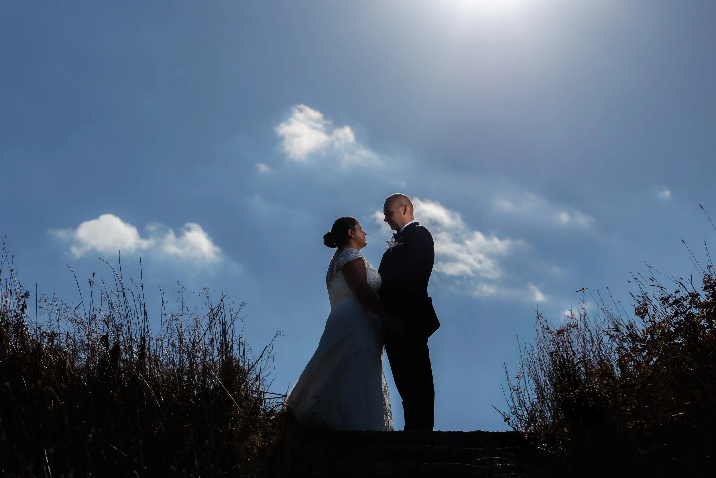 A bride and groom standing close together outdoors, holding hands and facing each other, with a bright sky and clouds in the background.