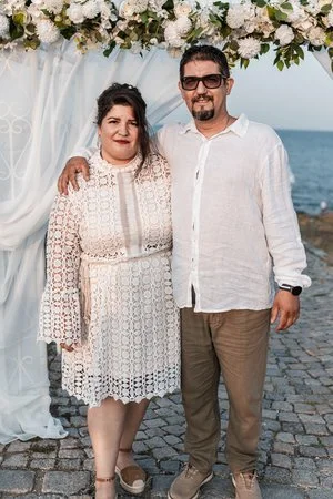 A man and woman standing together outdoors by the water, under a white floral archway, dressed in white.