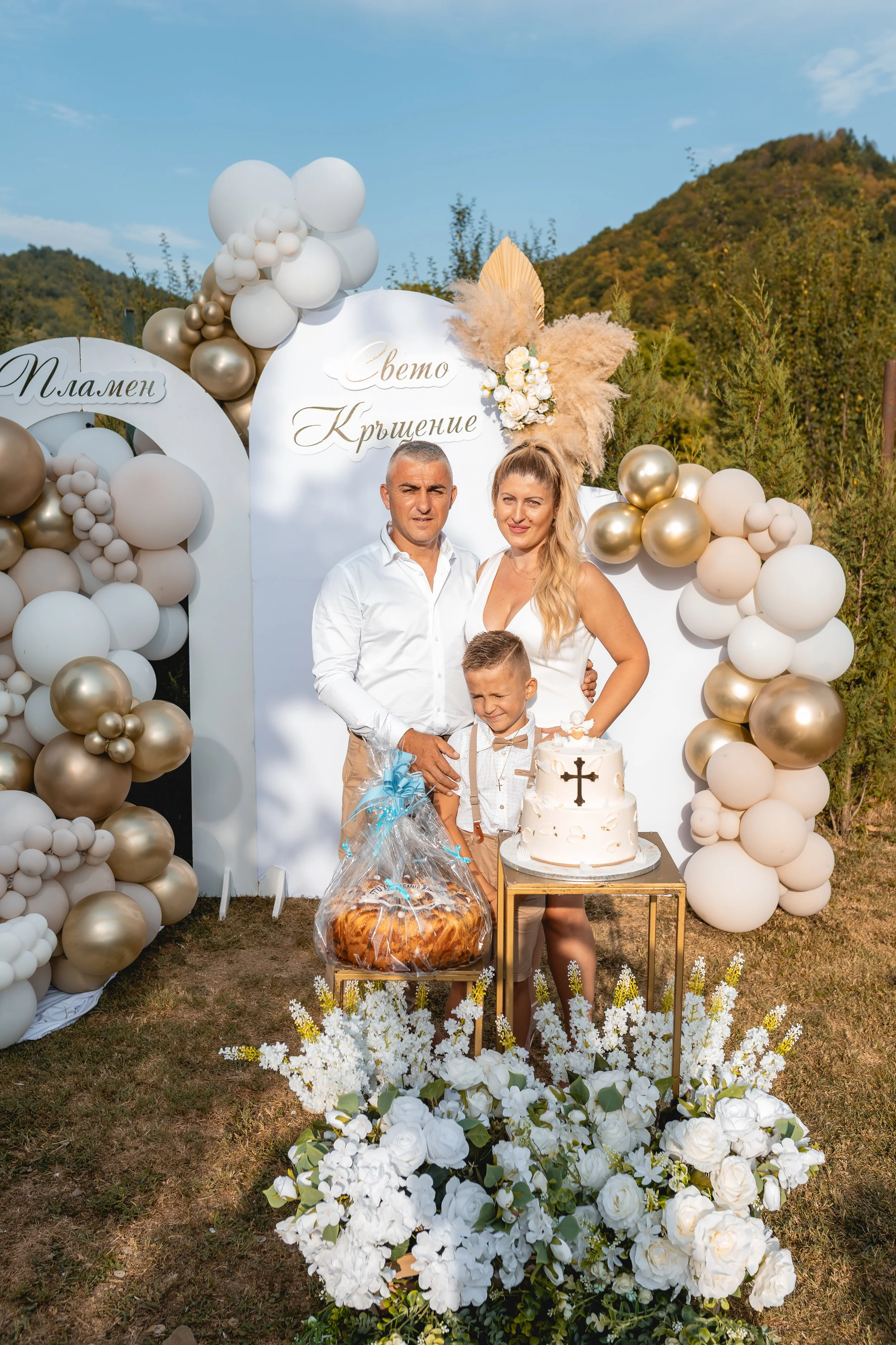 A family celebrates a religious ceremony outdoors with a decorated backdrop, a cake, a bread, and floral arrangements.
