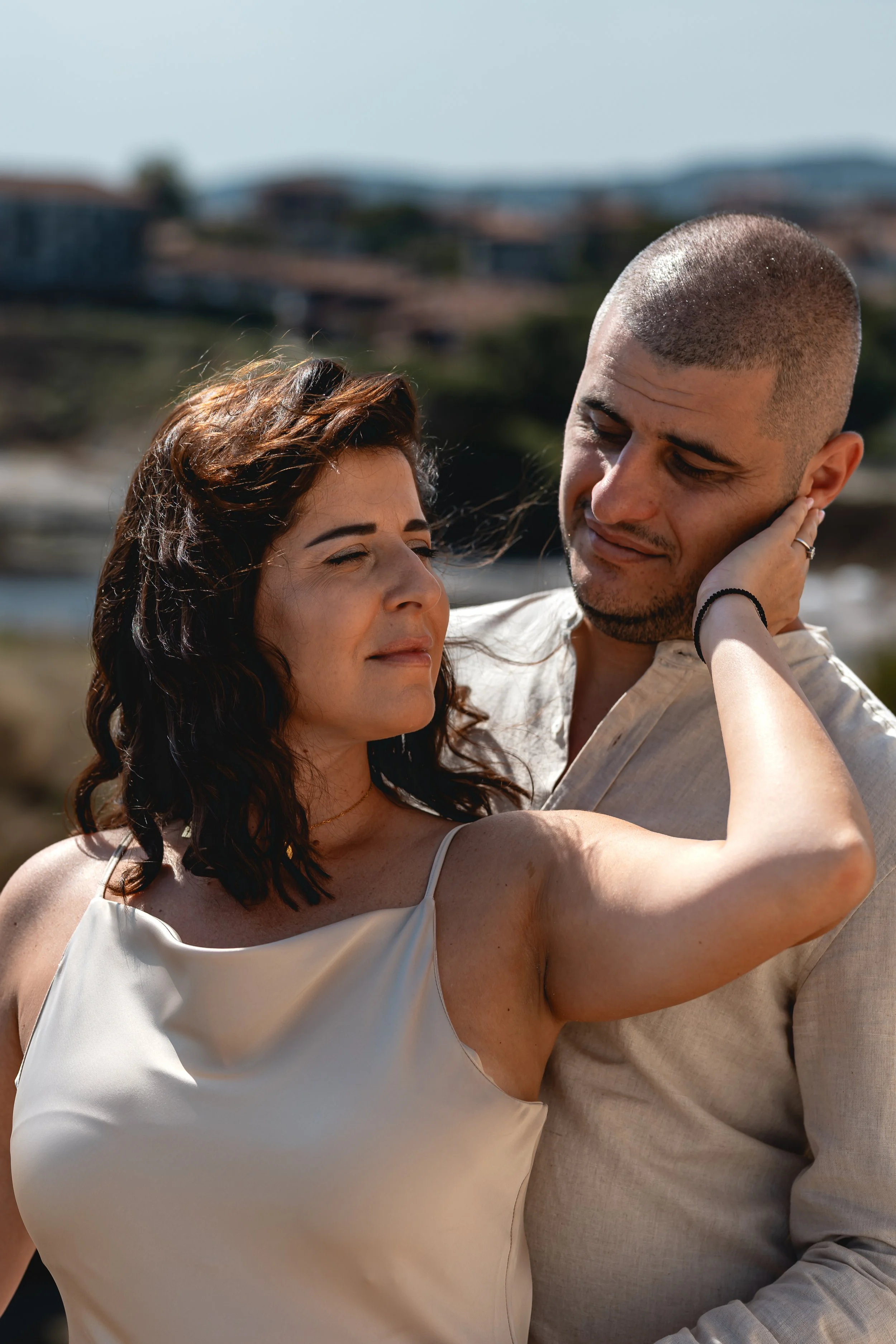A woman with dark wavy hair and a light-colored sleeveless top touching a man's face. The man has a shaved head and is wearing a light-colored shirt. They are outdoors with blurred houses and landscape in the background.