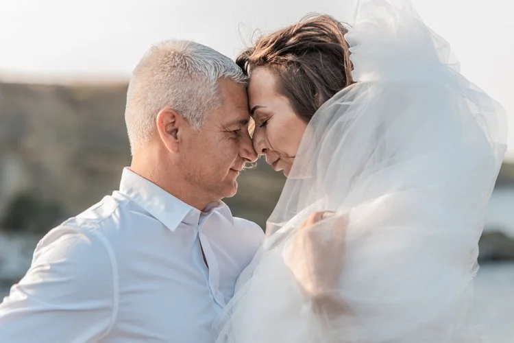 A couple with their foreheads and noses touching, dressed in wedding attire, outdoors near water.