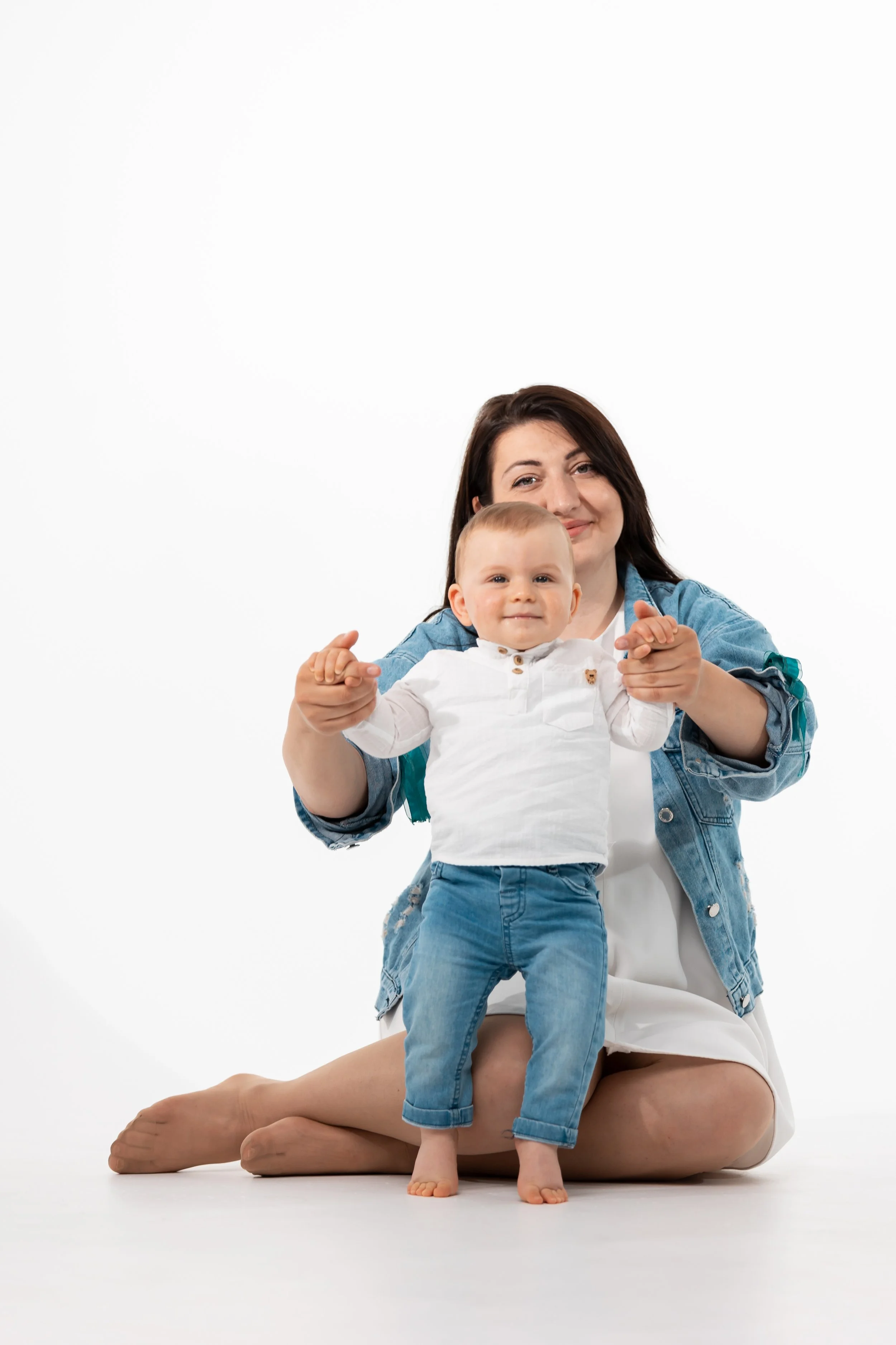 A woman kneeling on the floor, holding a young child by his hands, with both smiling at the camera against a plain white background.