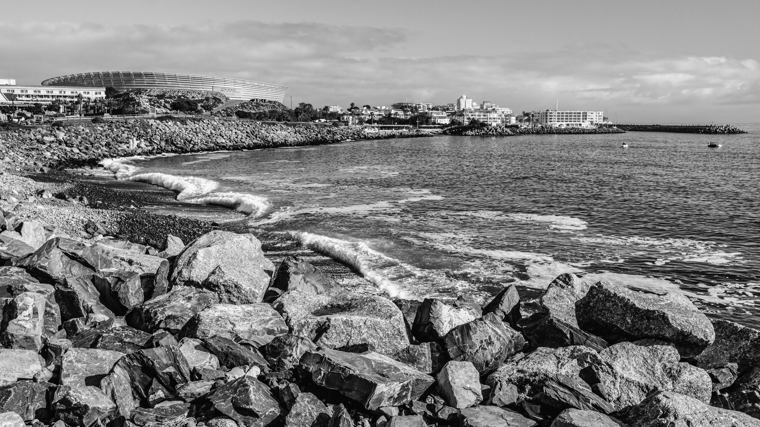 Black and white photo of a rocky shoreline with small waves, and a cityscape with buildings and a stadium in the background along the coast. Cape Town, South Africa.