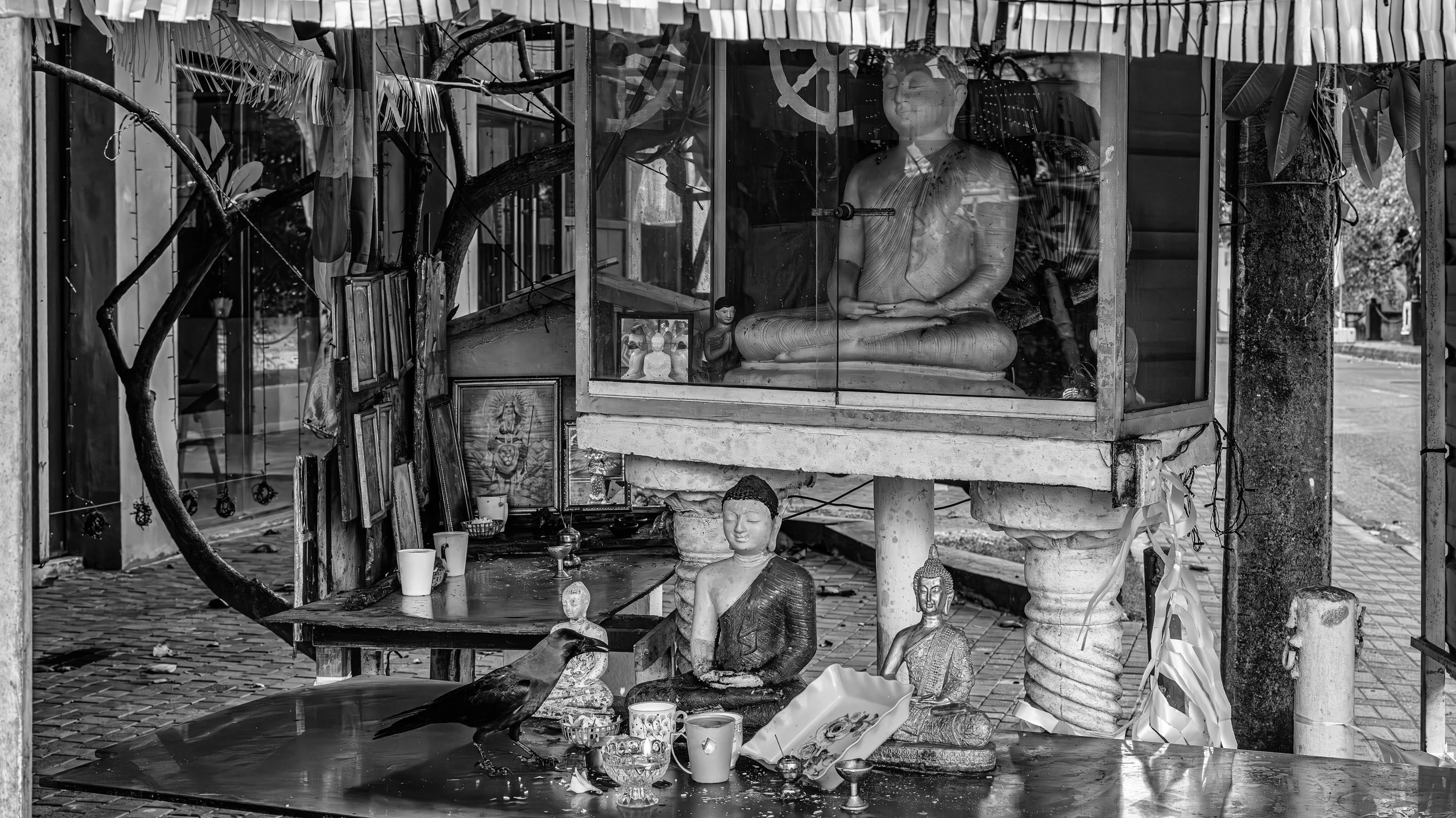 A storefront display featuring Buddha statues, framed religious artwork, and various small religious icons and offerings, with residential buildings and street visible in the background.