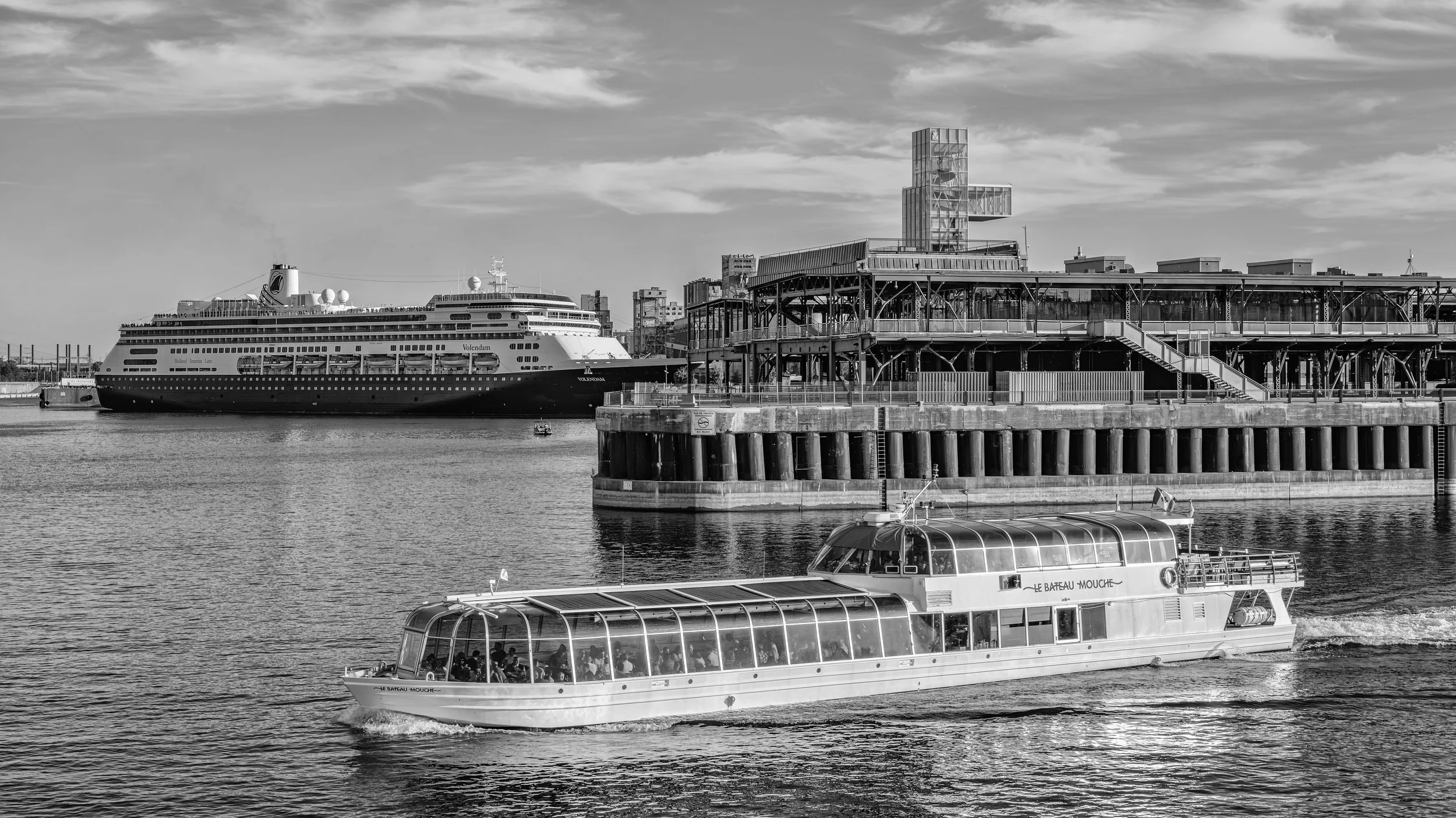 A harbor scene in black and white with a cruise ship docked, a ferry boat in motion, and a modern building with glass architecture against a cloudy sky.