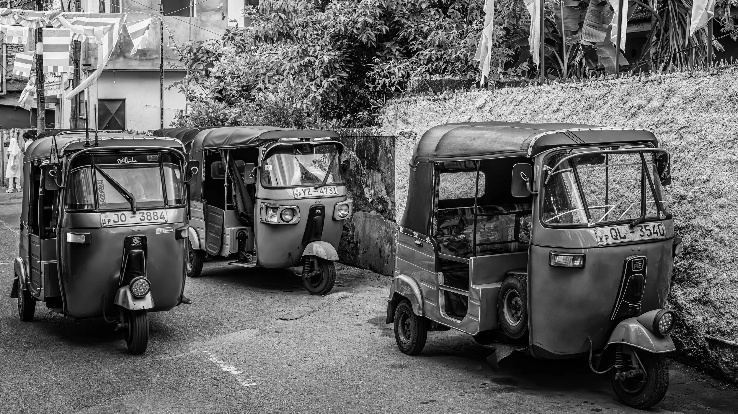Three auto rickshaws parked on a street next to a concrete wall, with plants and buildings in the background.