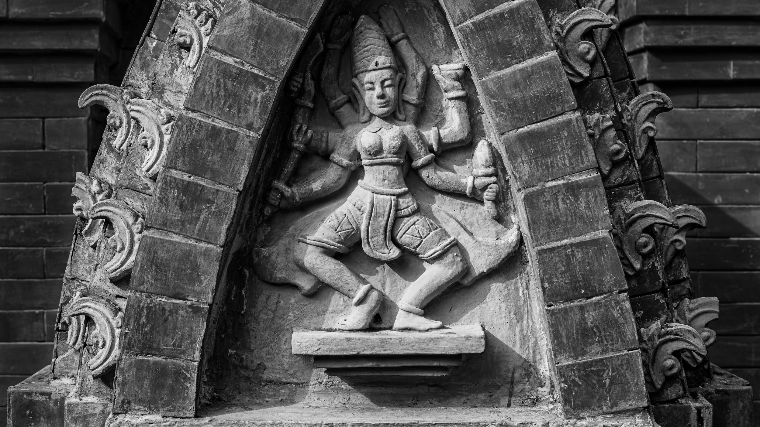 A carved stone religious figure, possibly a deity or saint, depicted in a dancing pose, surrounded by ornate decorative elements, set against a brick background.