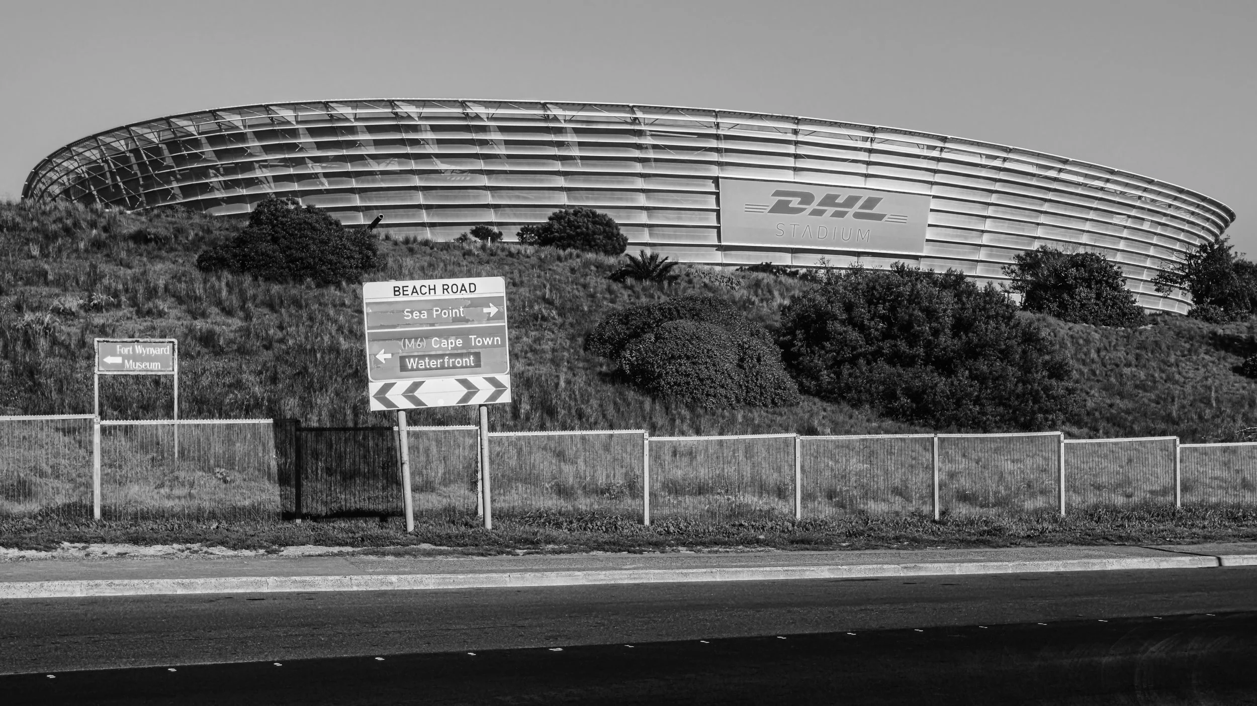 Black and white photo of a modern stadium labeled 'DHL Stadium' on a hillside with road signs pointing to Beach Road, Sea Point, Cape Town, and Waterfront. Cape Town, South Africa.