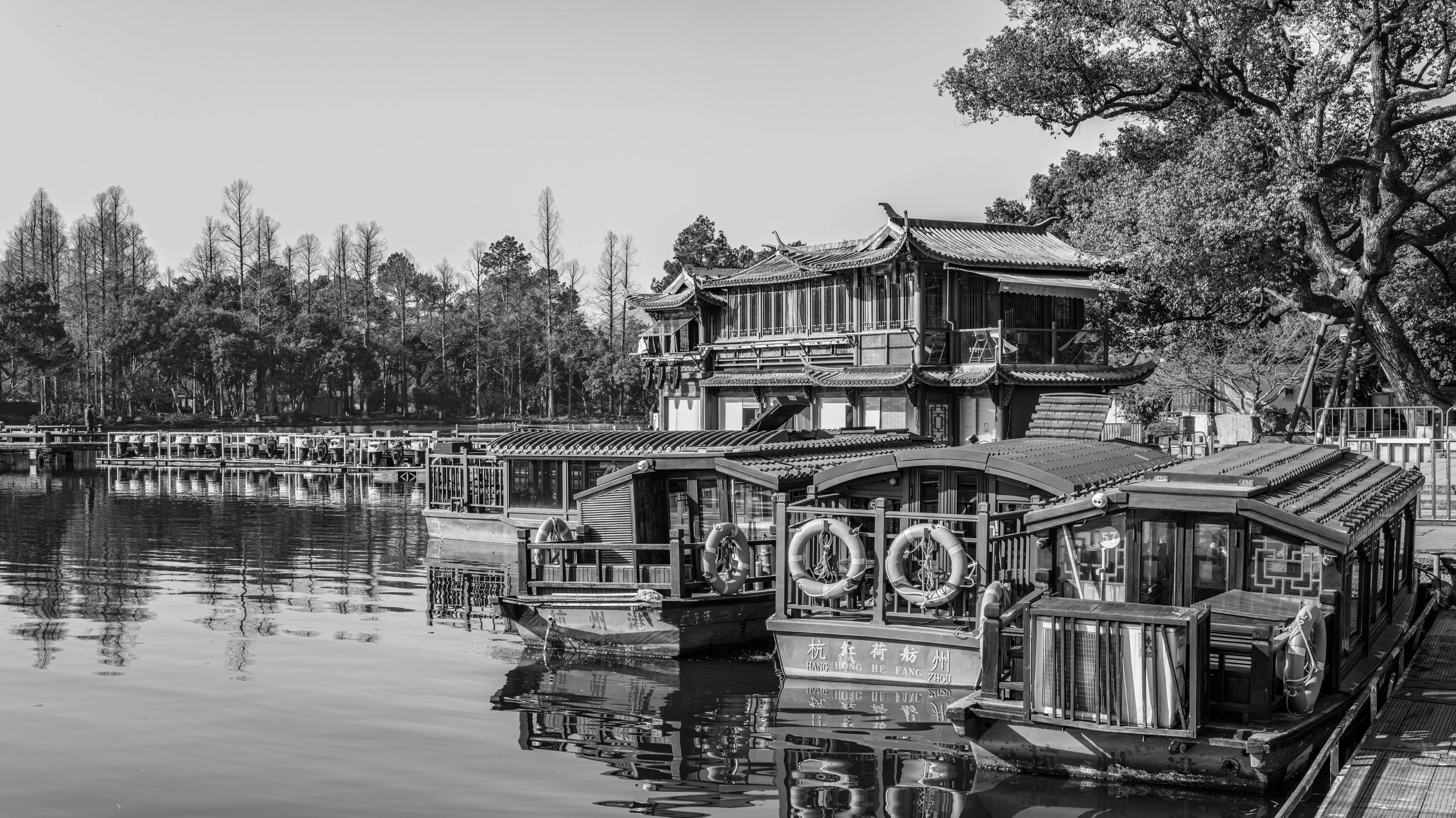 A black and white photo of traditional Chinese architectural buildings on a waterfront with trees in the background.