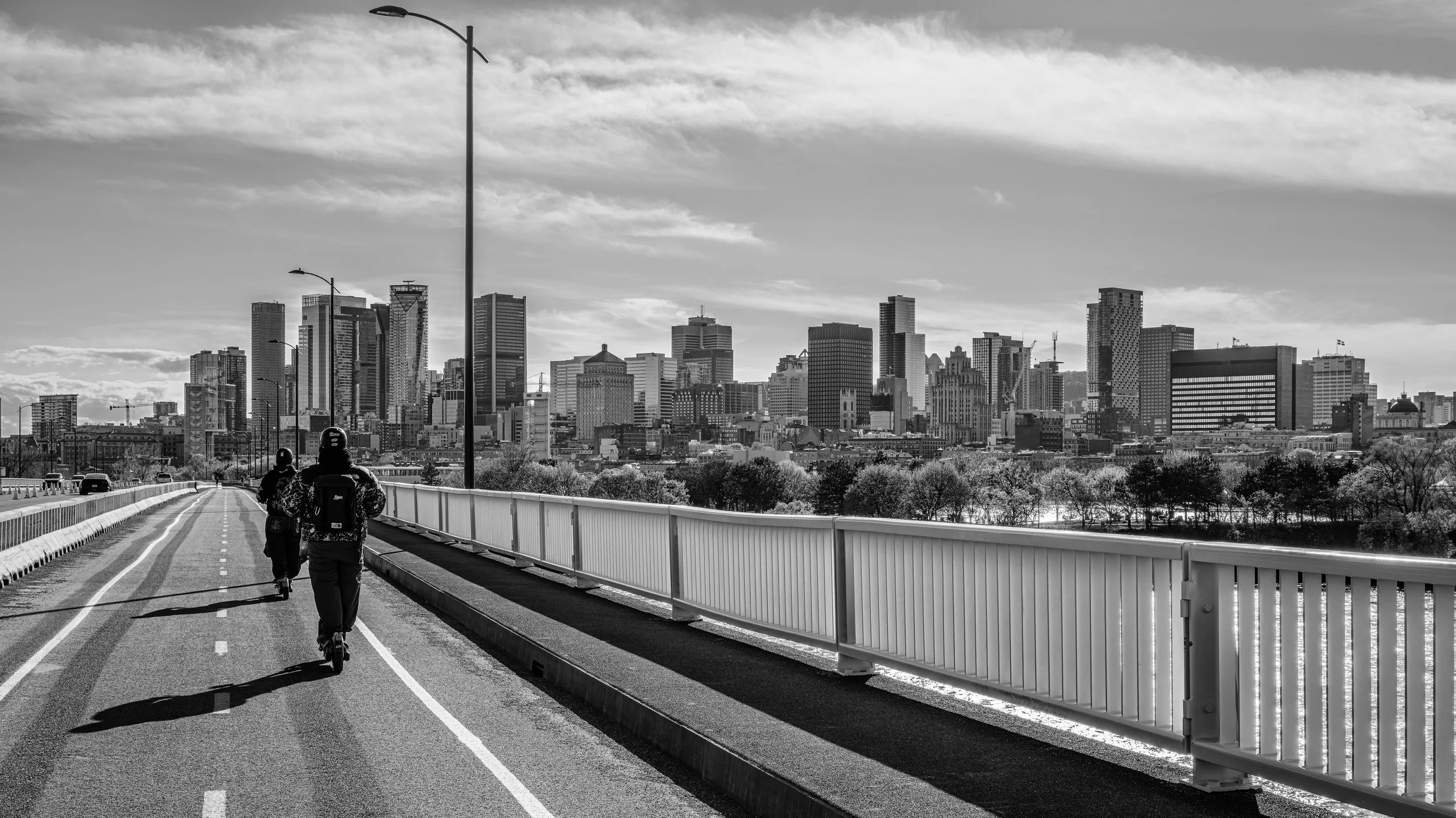 Two cyclists riding on a paved multi-lane road with a city skyline in the background. The scene is in black and white.