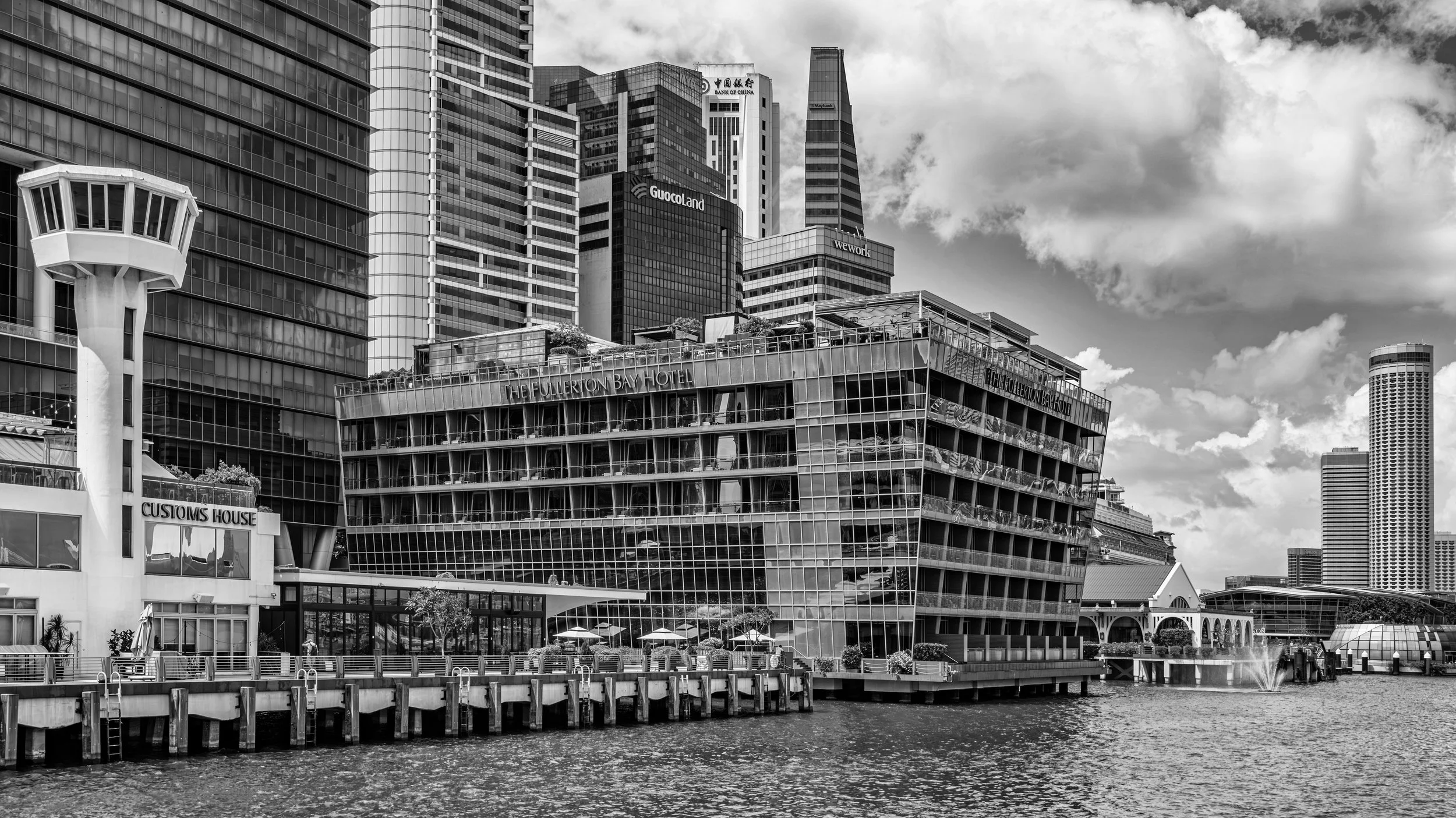 Black and white photograph of modern city buildings along a waterfront, including the Fullerton Bay Hotel and other high-rise office buildings, with puffy clouds in the sky and a waterway in the foreground.