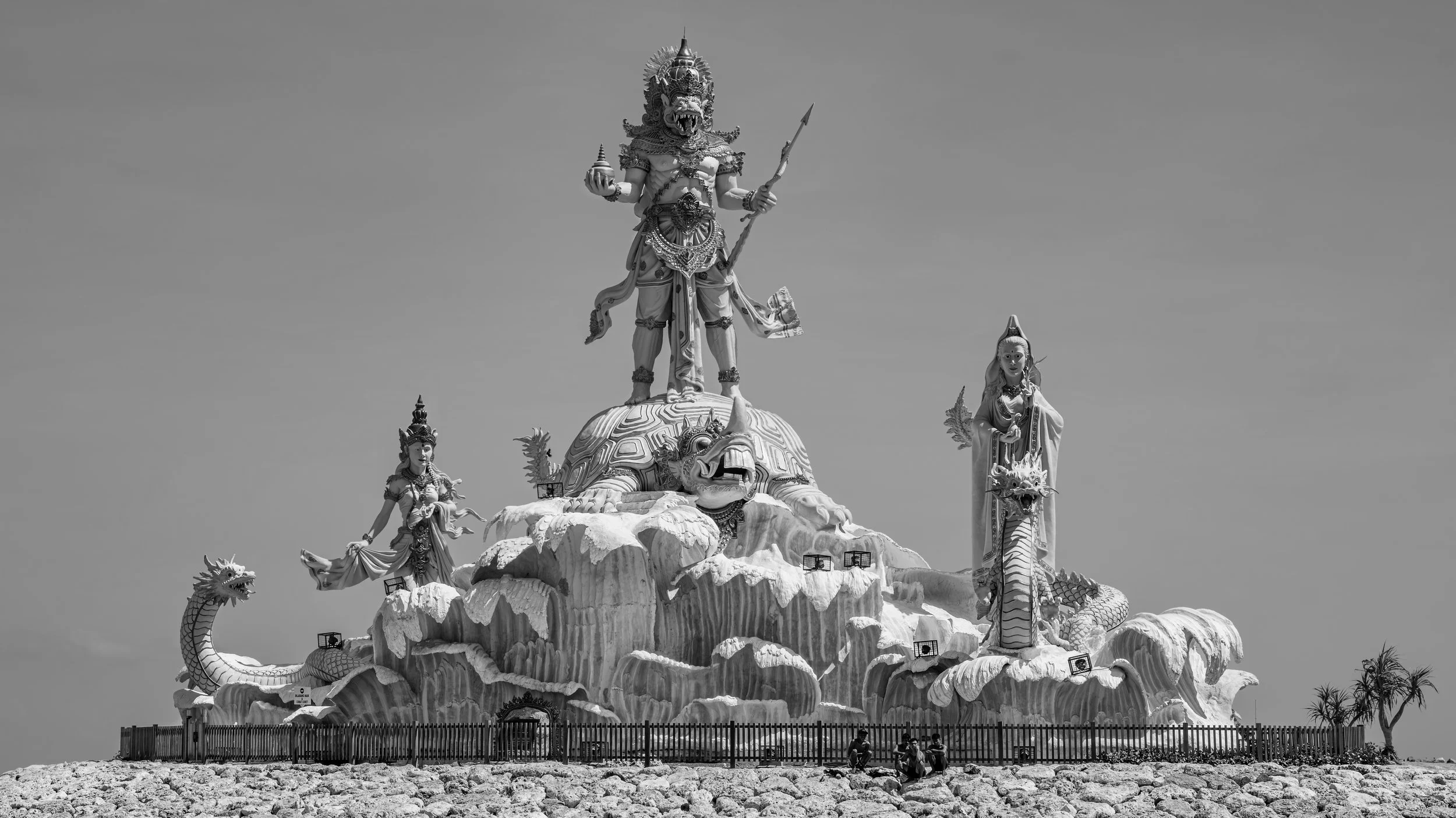 A large elaborate monument featuring mythological figures including a central deity with a fierce lion-like face, holding a sphere and a spear, surrounded by women, dragons, and a turtle base, enclosed by a fence, with people at the bottom.