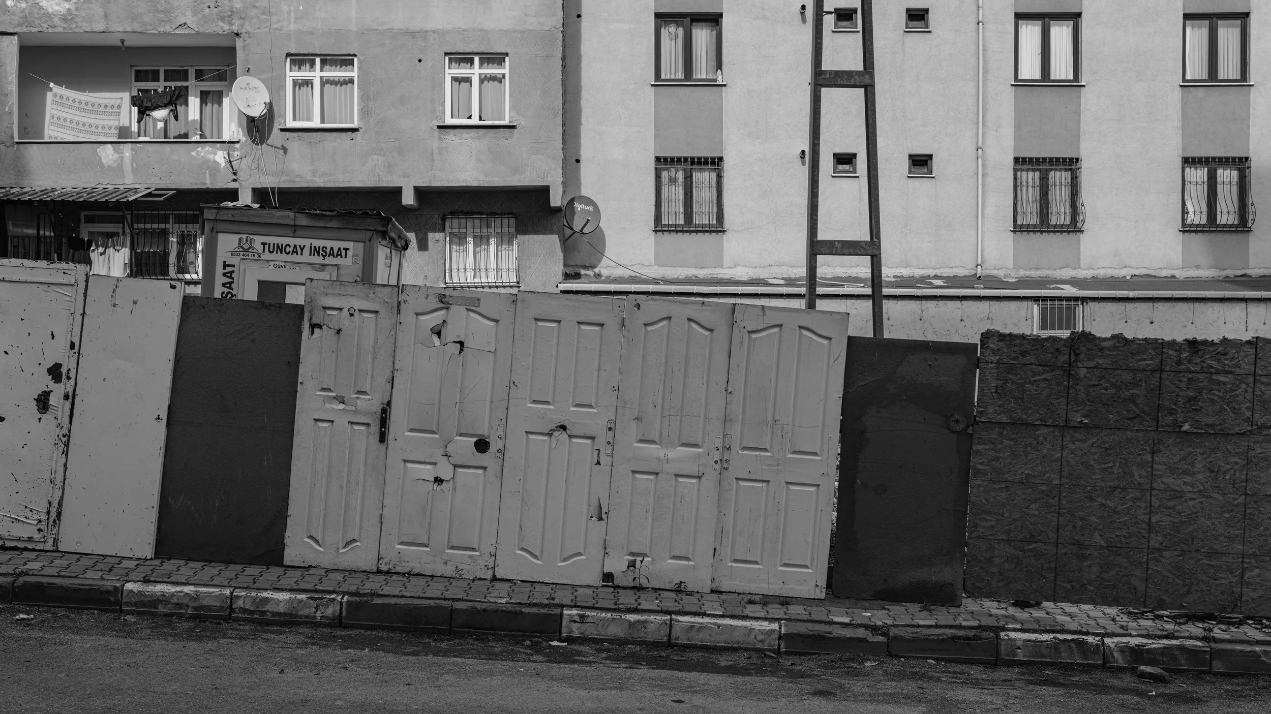 A black and white photo of an urban scene with torn and weathered doors leaning against a low wall, on the street in front of a multi-story residential building with several windows, satellite dishes, and laundry hanging outside one window.