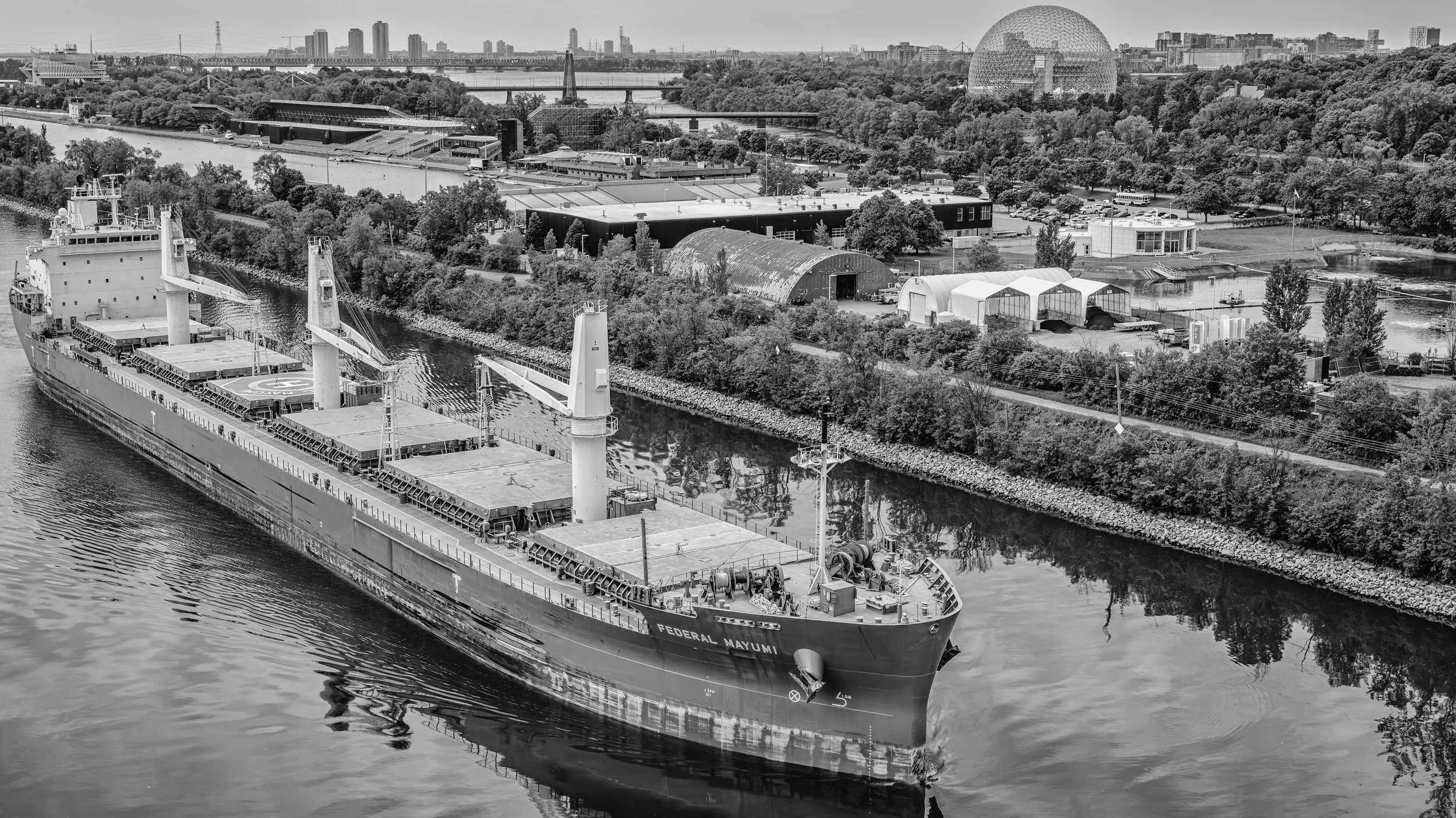 Aerial view of a city skyline with a river, a cargo ship named Federal Mayumi in the foreground, green trees, and modern buildings in the background, in black and white.