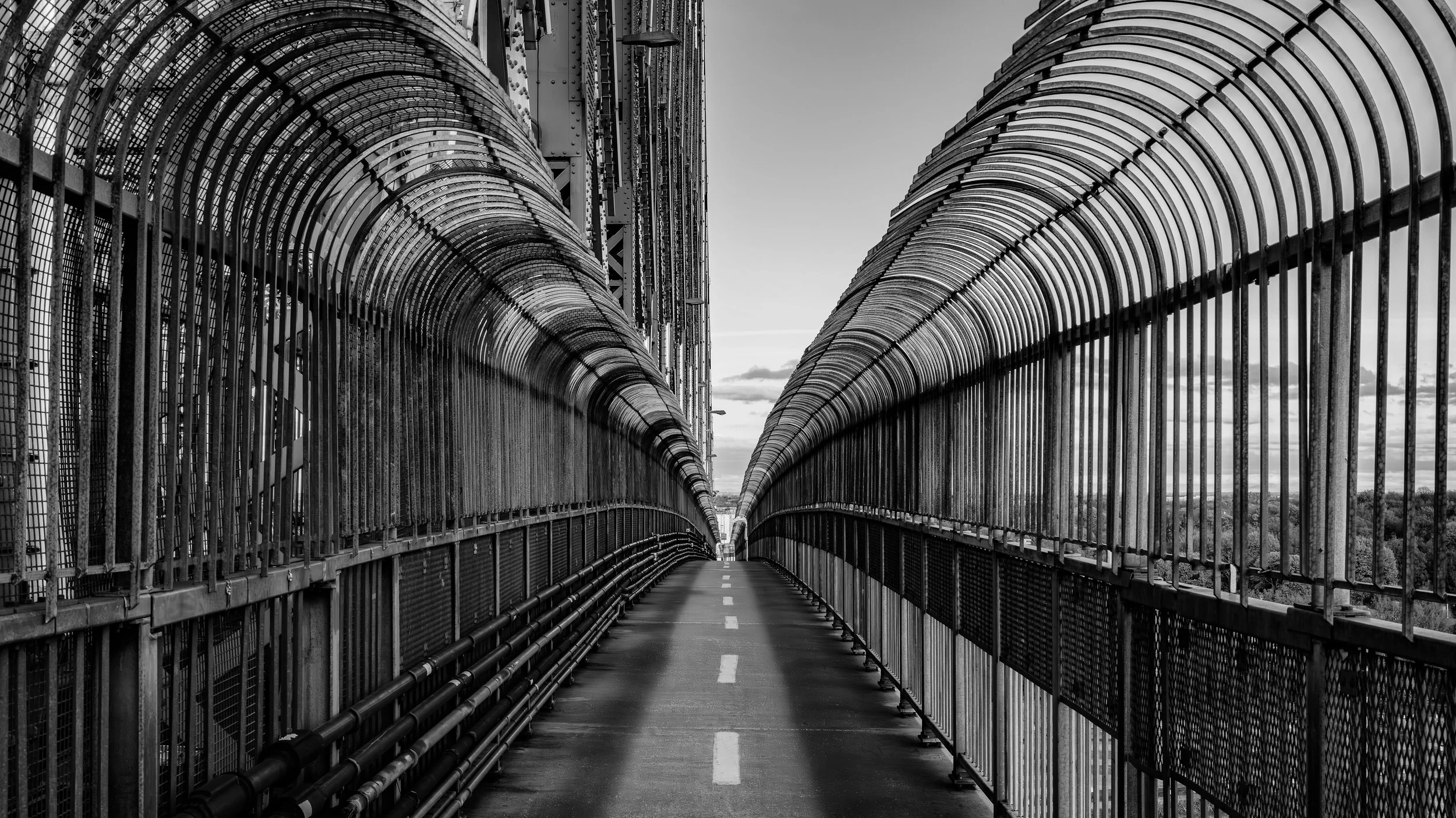 Black and white photo of an enclosed pedestrian bridge with metal fencing on both sides, curving to the left, with a dashed white line down the center. The sky is cloudy in the background.