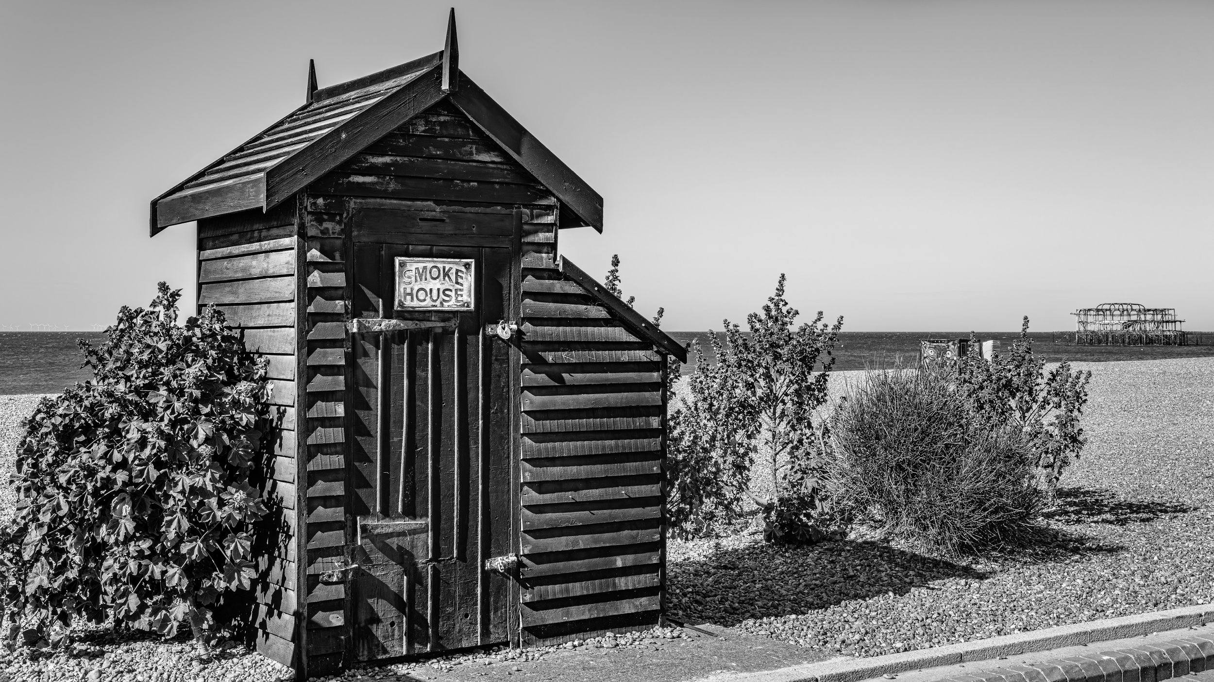 Black and white photo of a small wooden beach hut with a sign that reads 'Smoke House,' located on a pebble beach with some plants and bushes around, and the ocean in the background along with a pier or structure extending into the water.