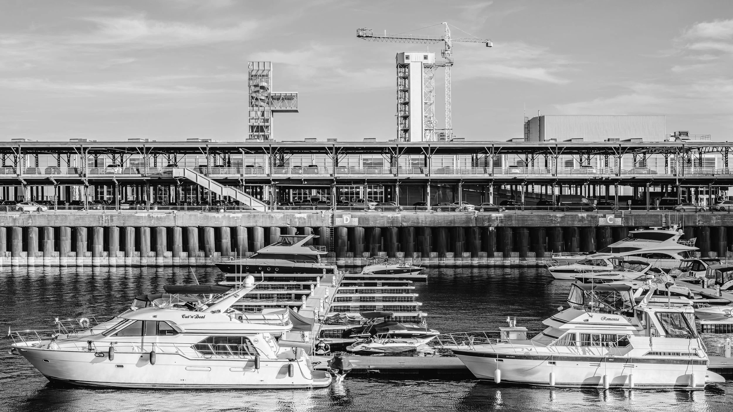 Black and white photo of a harbor with boats docked in the water, a multi-level structure with cars on top, and a construction crane in the background.