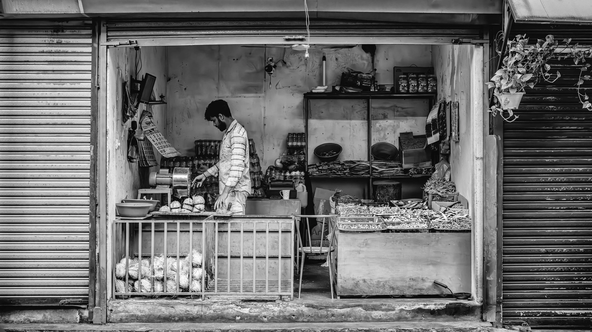 A man working at a small street shop selling snacks and other items, with a variety of goods displayed in front and on shelves inside, in a black and white photograph.