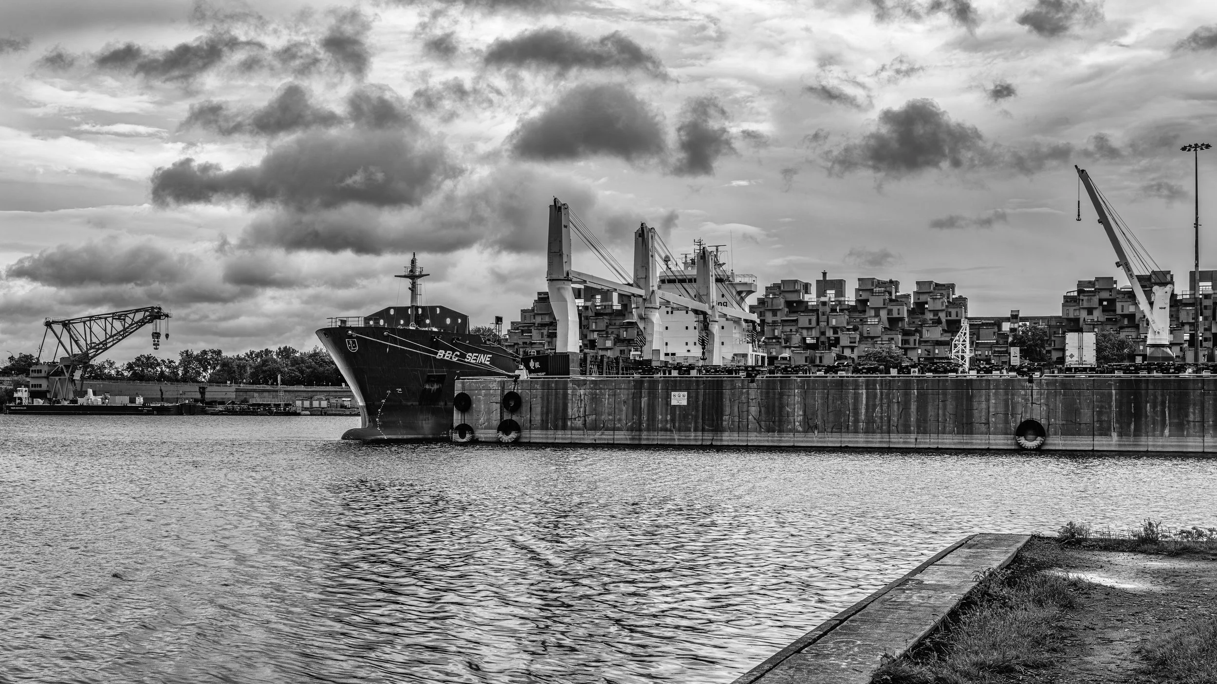A black and white photo of a docked cargo ship named BBC Seine, with large cranes on the deck, along a waterfront with buildings and cloudy sky in the background.