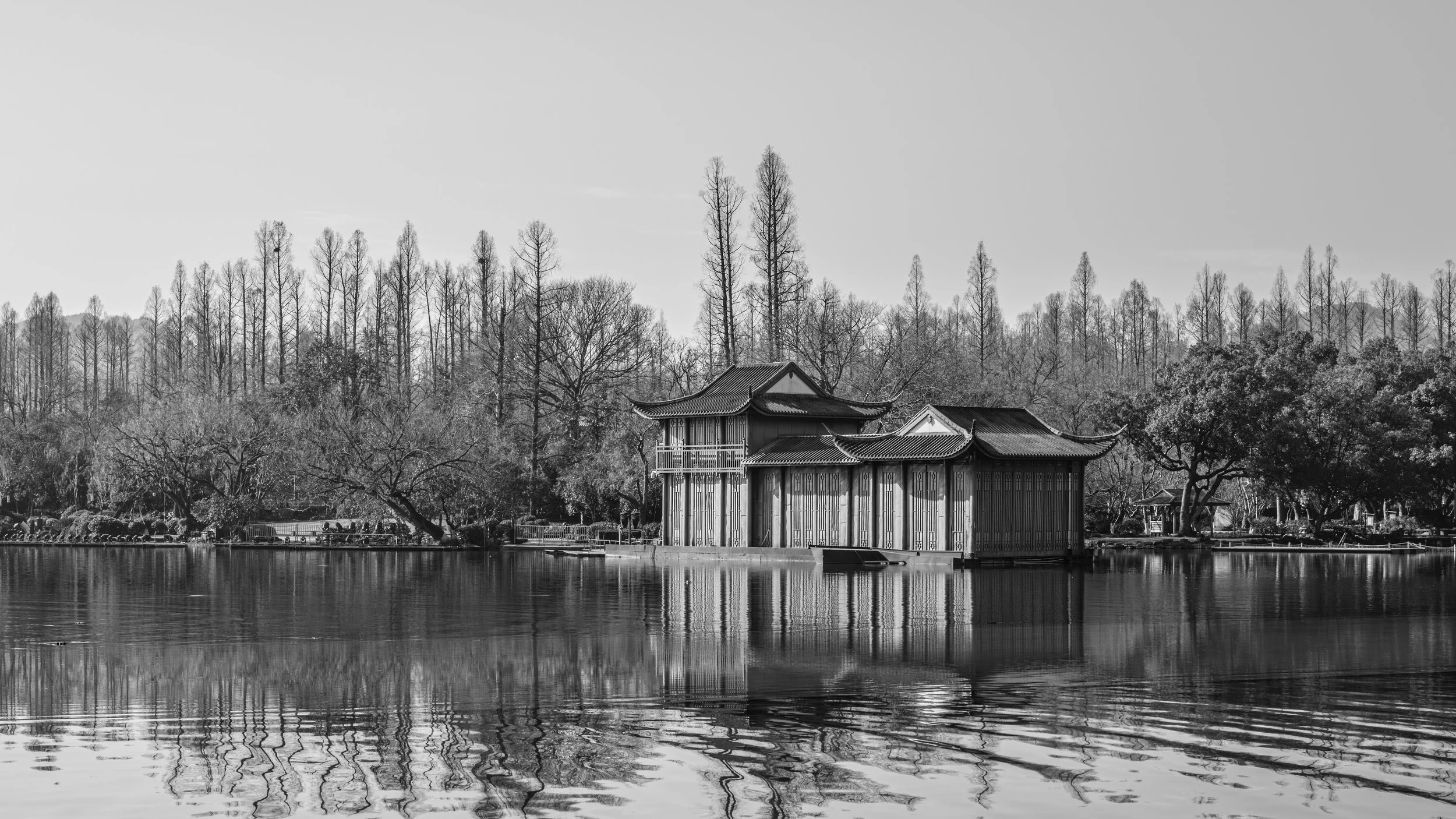 Black and white photo of traditional Asian-style buildings by a water body, with tall trees in the background.