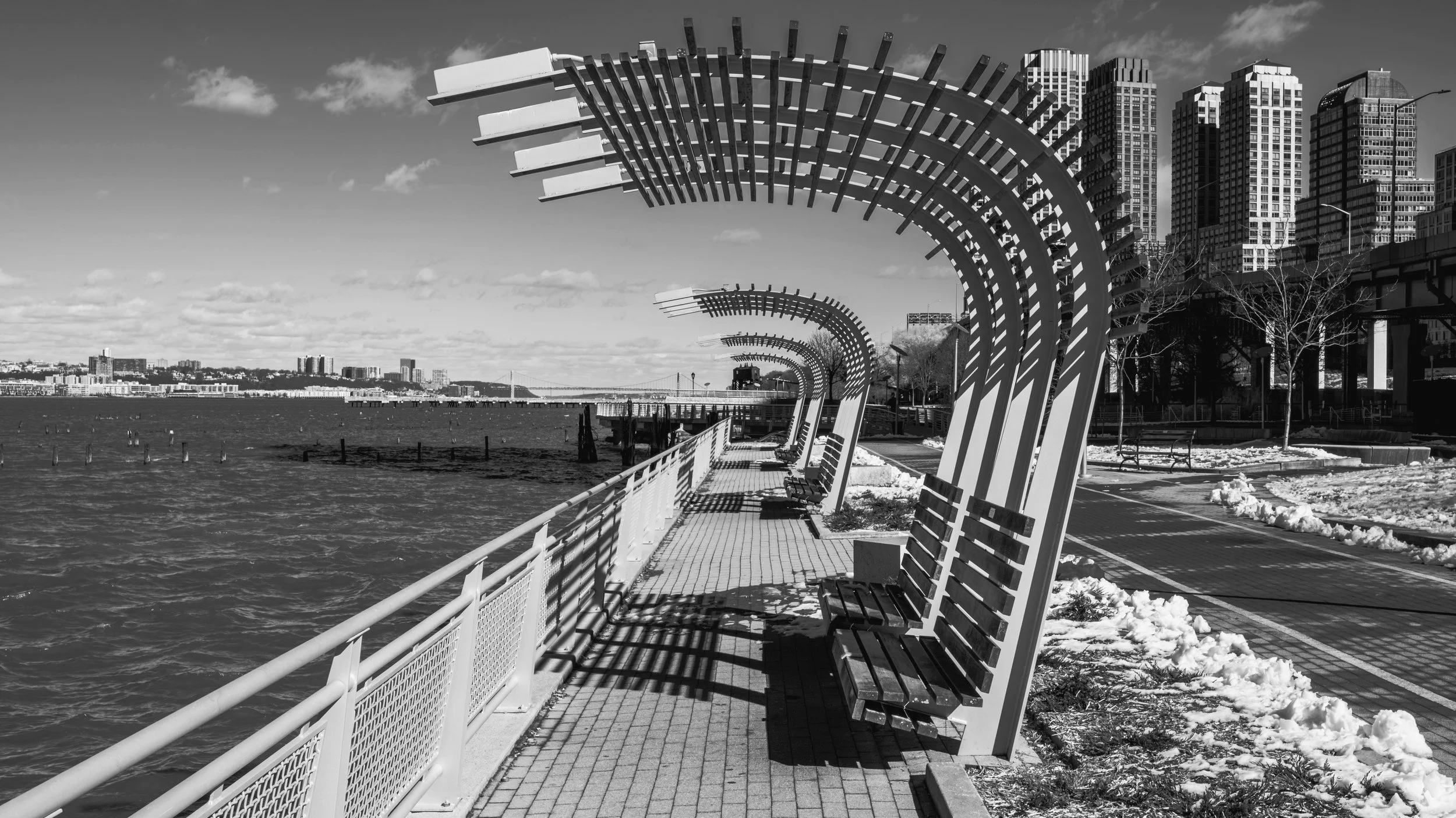 Row of outdoor benches along a waterfront promenade with modern city skyscrapers in the background, in black and white. Manhattan, New York, USA.