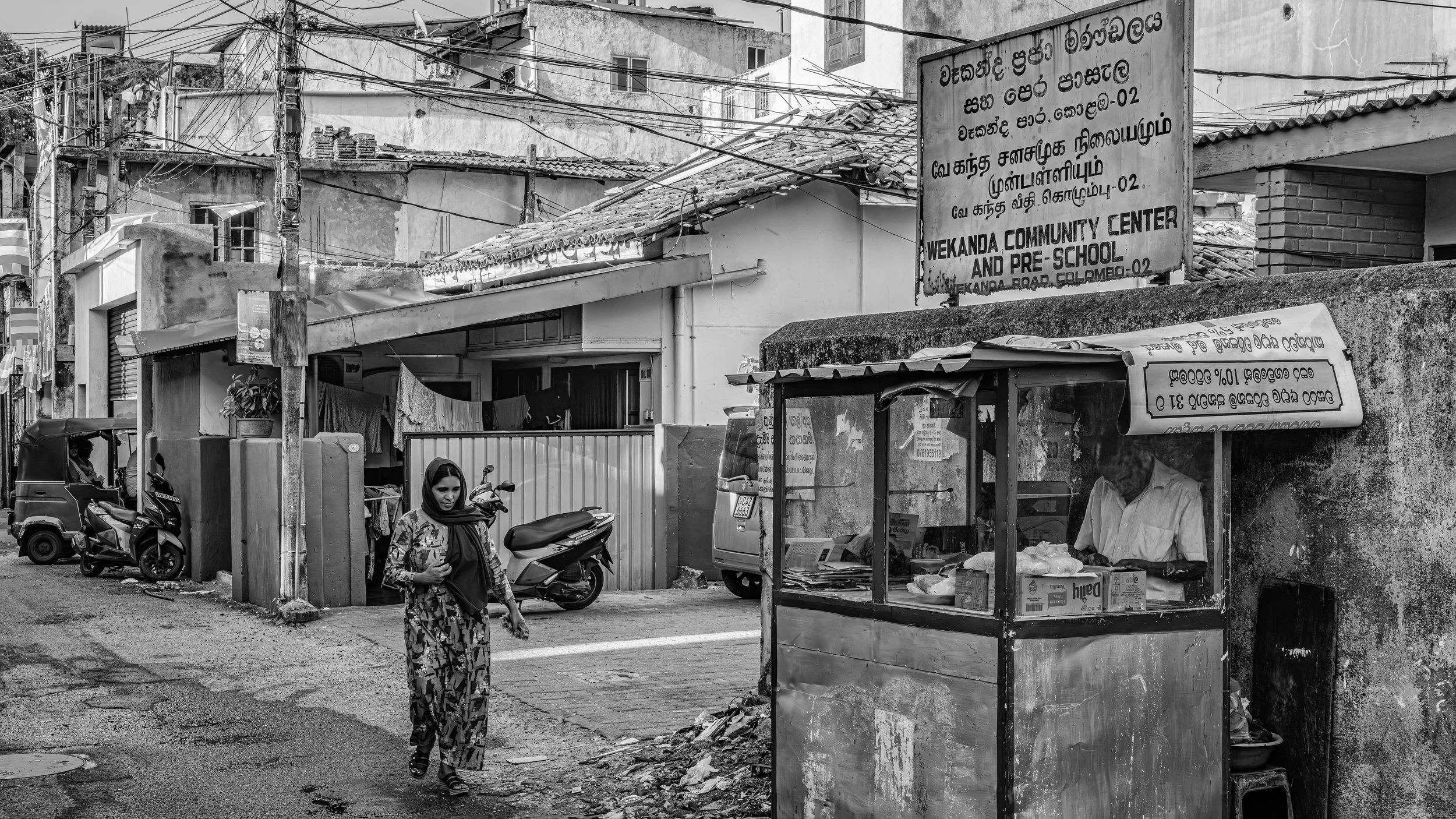 Street scene in an urban area with buildings, power lines, and a bus stop. A woman in patterned clothing and head covering is walking, and a man is working at a small roadside shop. Signage in a local language and English advertises the Wekanda Commu