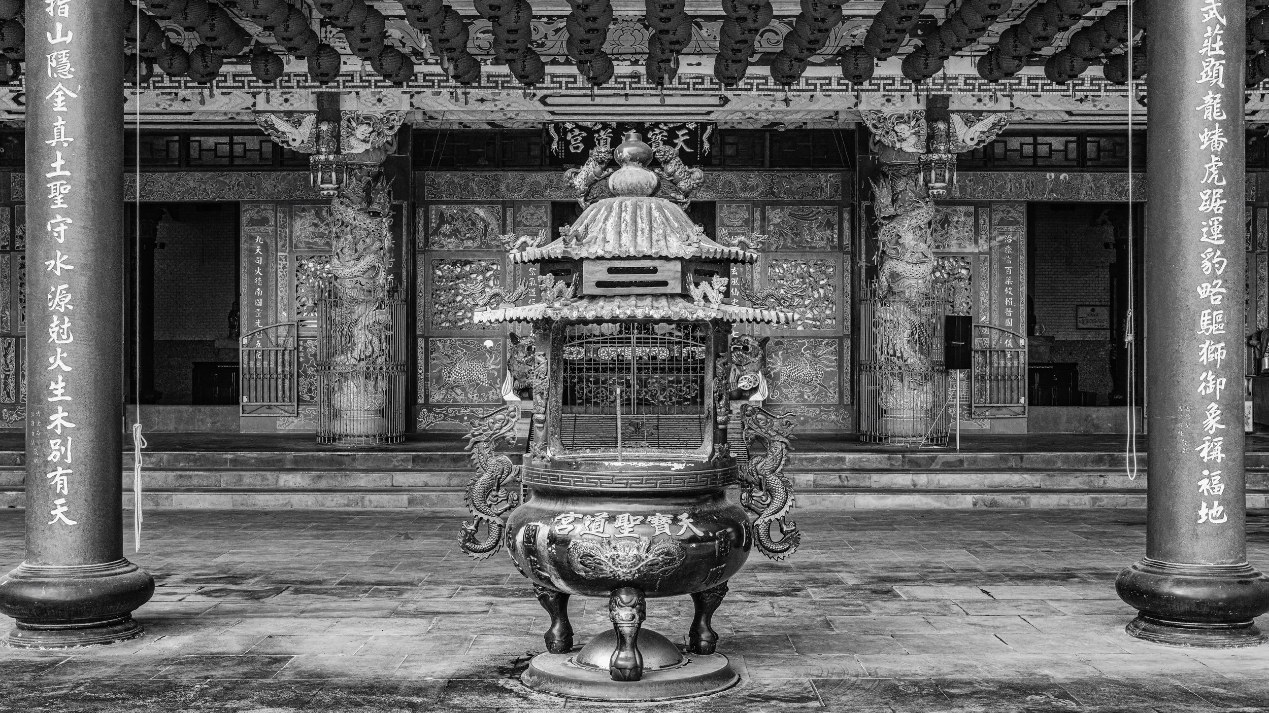 Black and white photo of an ornate temple interior with a central incense burner flanked by dragons, backlit wooden carvings, and large columns with Chinese characters.