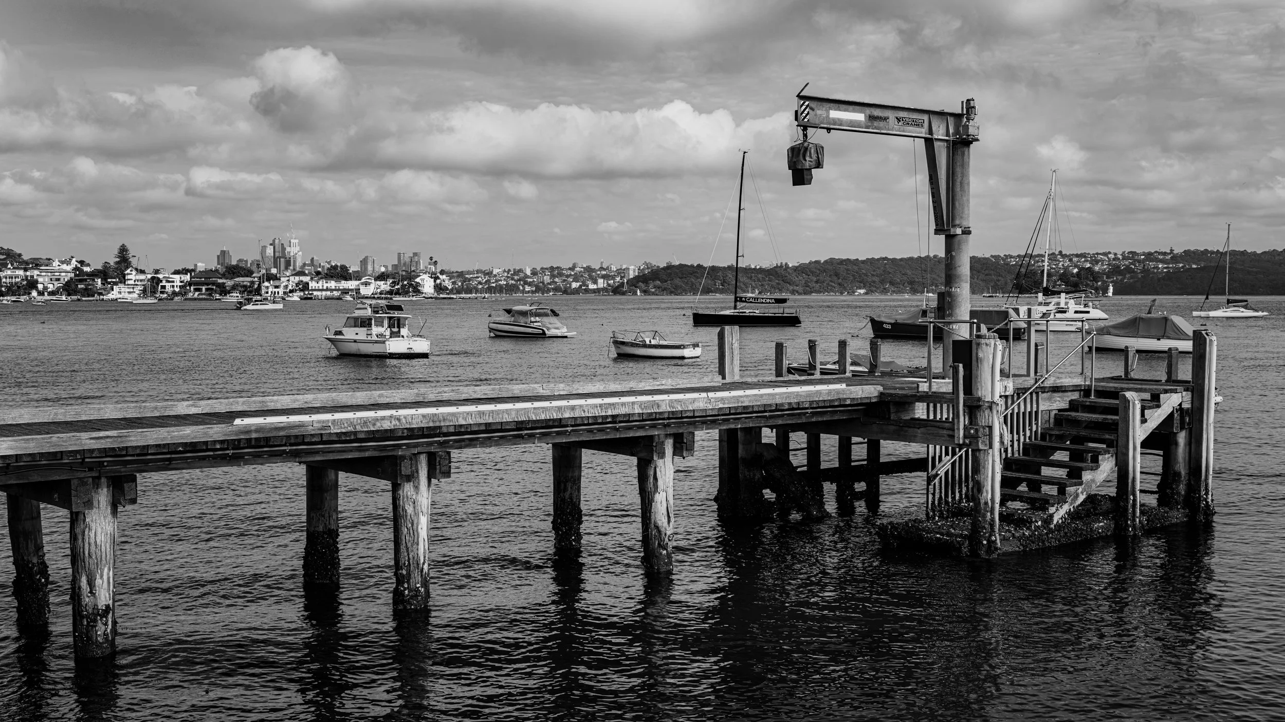 A black and white photo of a dock with boats moored in the water, a crane on the dock, and a city skyline with high-rise buildings in the background under a partly cloudy sky.