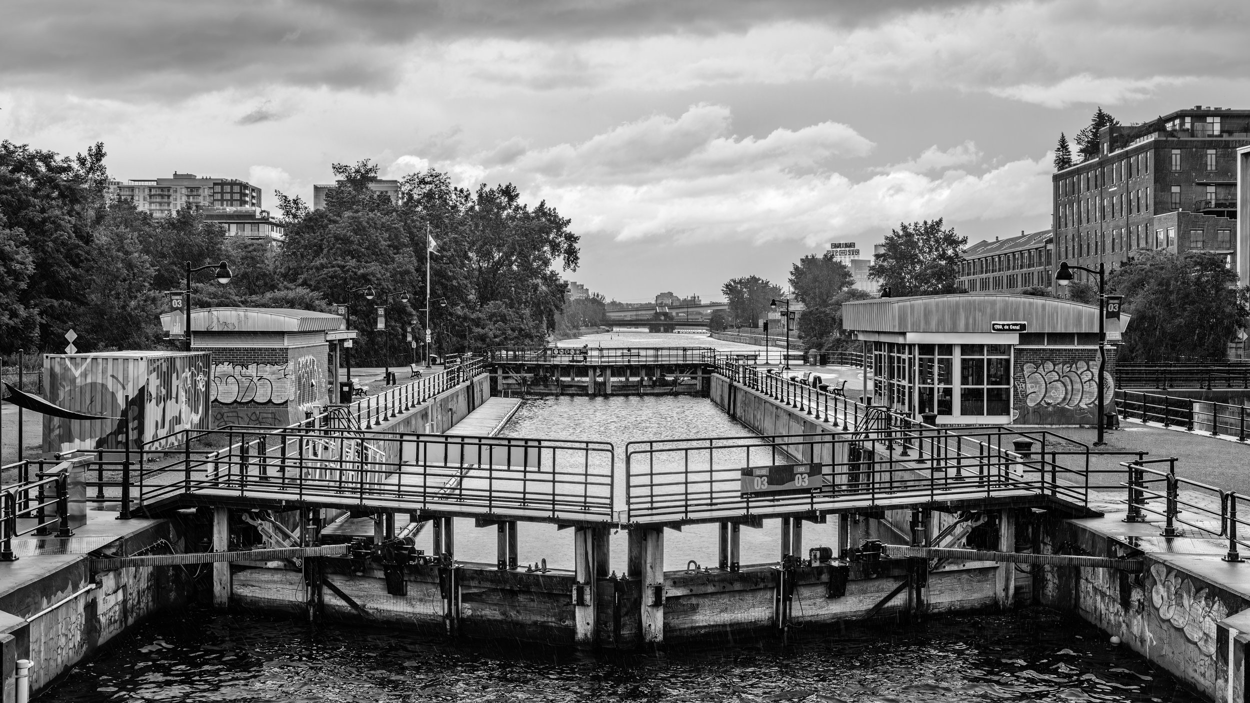 Black and white photo of a canal lock with infrastructure, graffiti on walls, and surrounding buildings and trees.