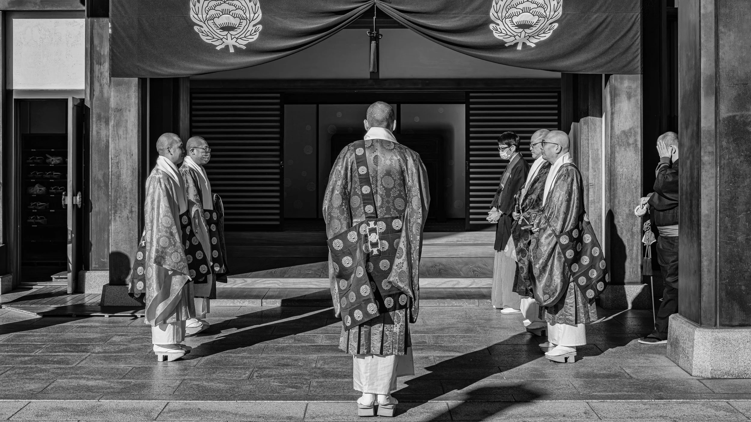 A group of men in traditional Japanese robes and masks stand in formation outside a building with wooden sliding doors, under fabric banners with floral emblems. Narita, Japan.
