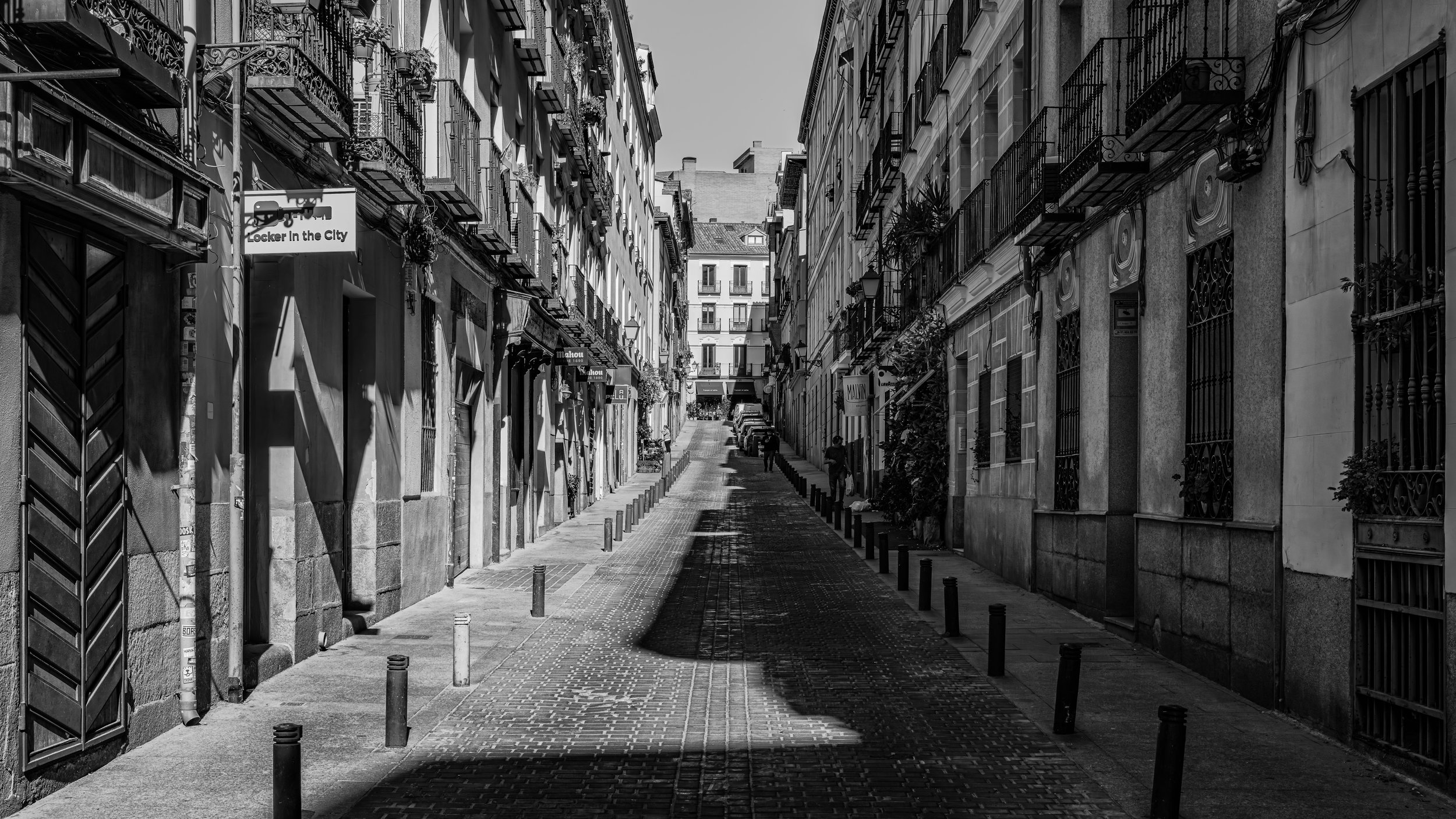 A narrow city street with tall buildings on both sides, featuring balconies with railings, some with plants. The street has bollards along the sidewalk, and a few pedestrians are visible in the distance. The image is in black and white.