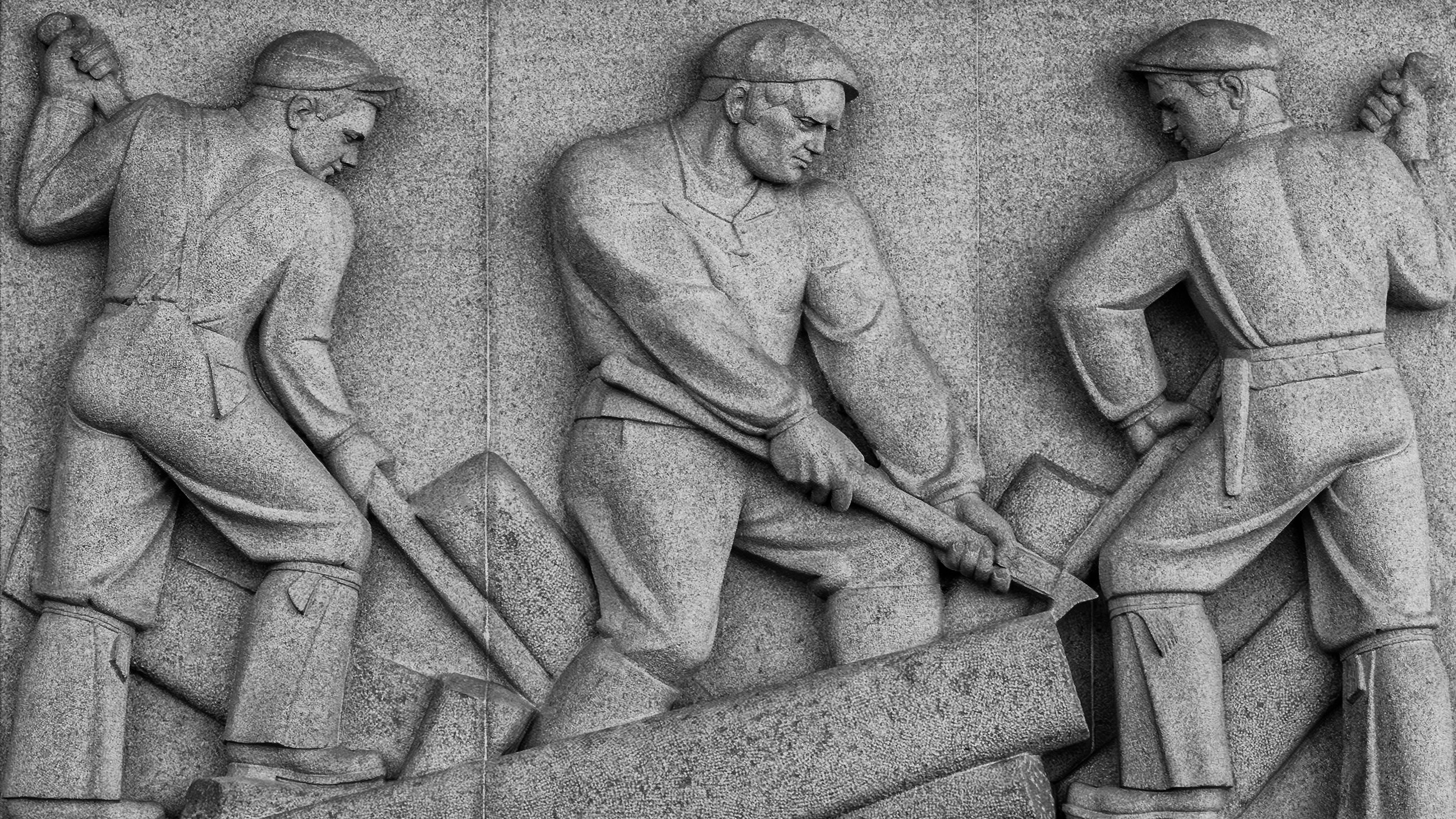 A black and white stone relief sculpture depicting three workers with pickaxes, involved in a labor-intensive activity.