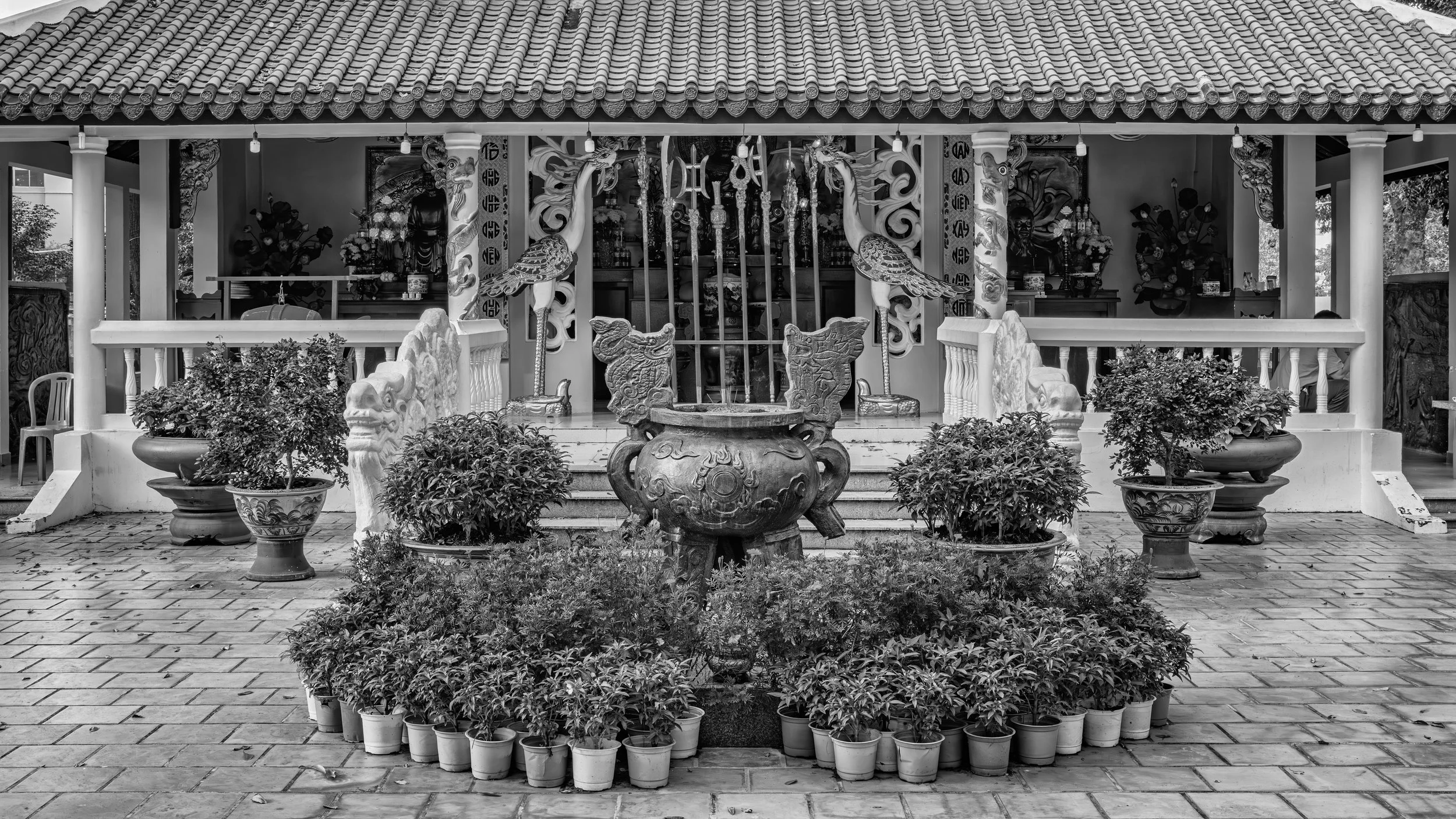 A traditional Asian temple with a tiled roof, ornate decorations, and surrounded by potted plants, in black and white.