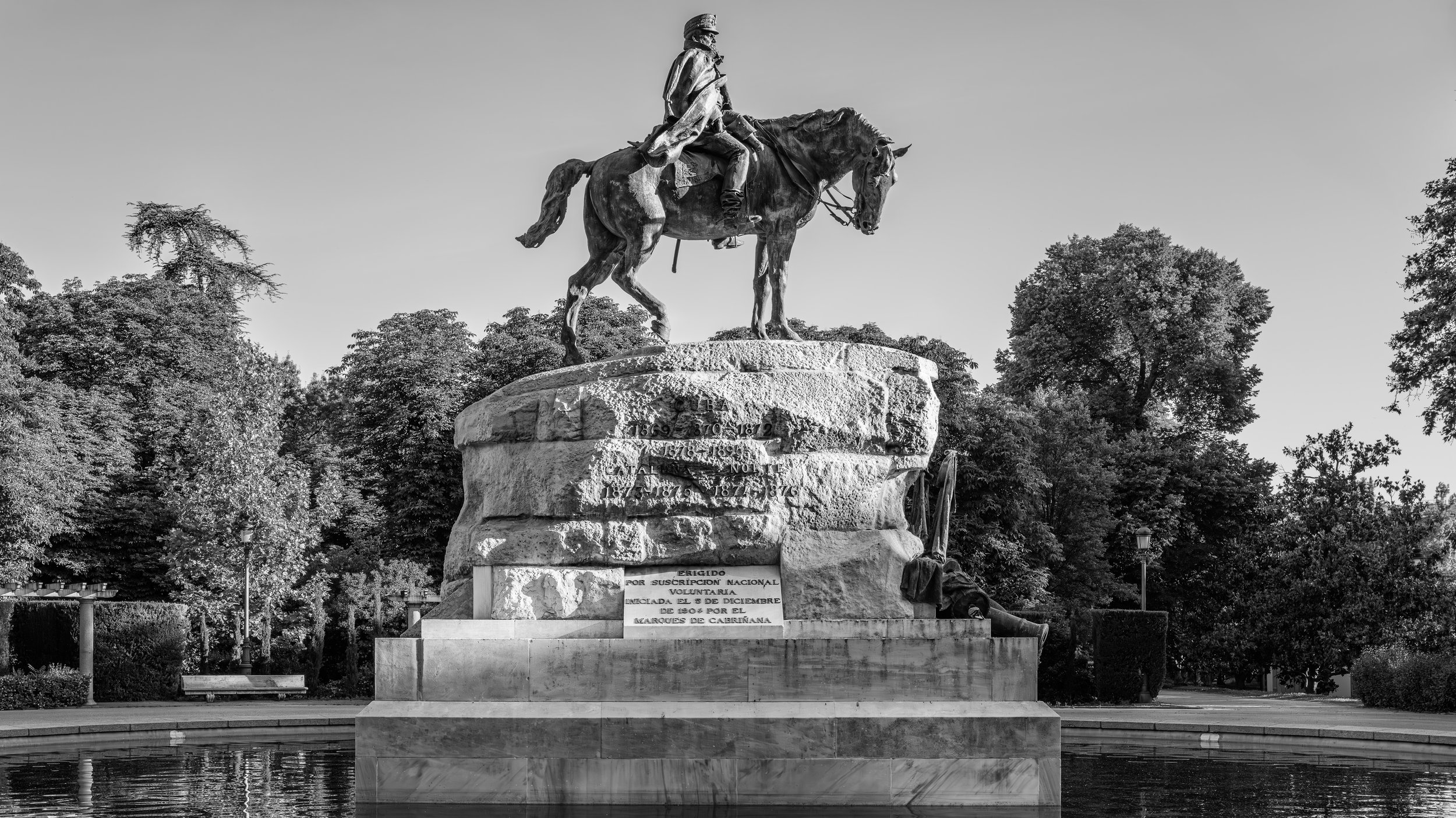 Black and white photo of a large equestrian statue featuring a man in a military uniform on a horse, mounted on a stone pedestal surrounded by trees and a reflective pool.