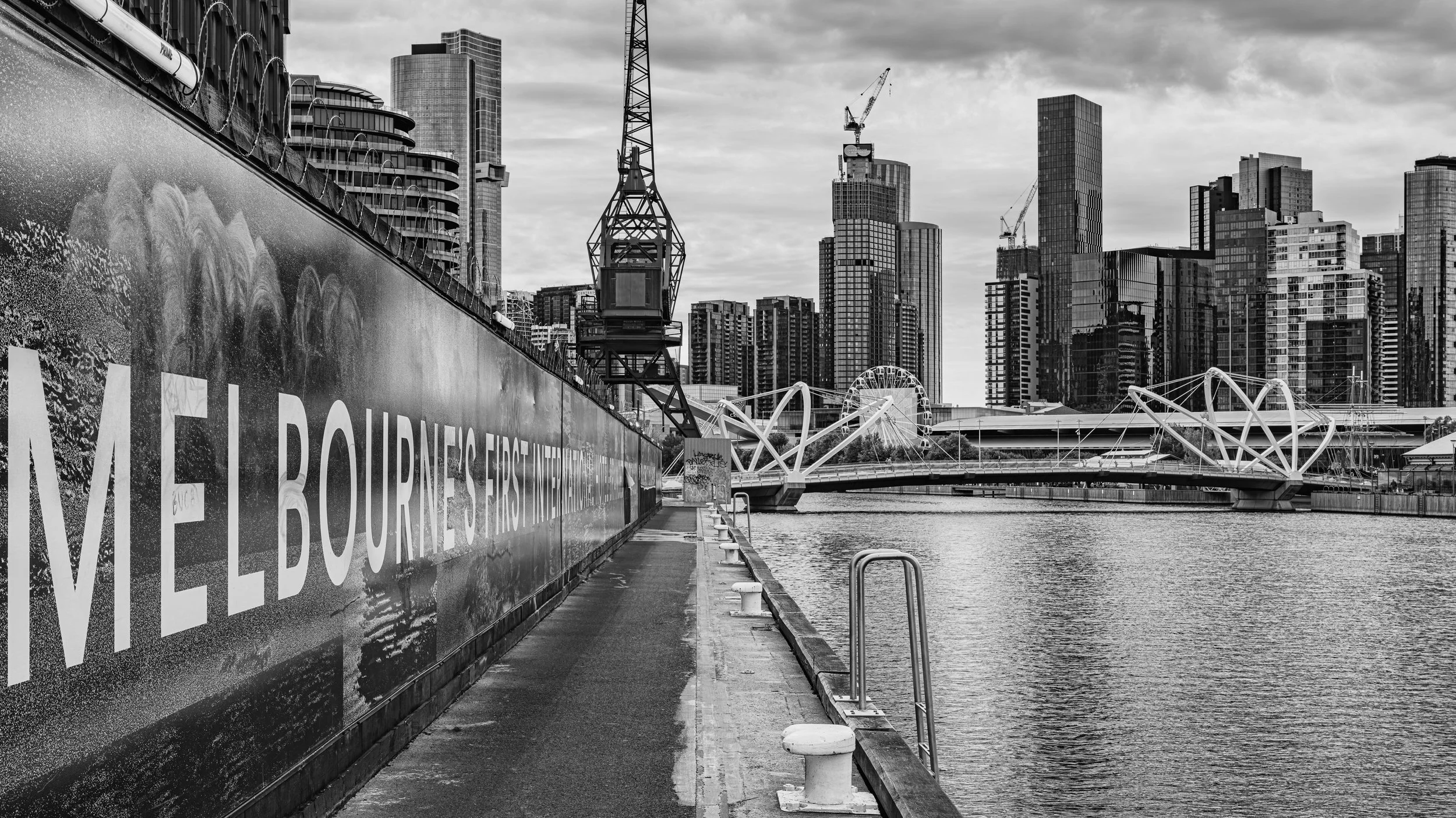 A black and white photo of a city skyline along a river, with high-rise buildings, a construction crane, and a modern bridge with white geometric arches. A walkway runs beside the water. Melbourne, Victoria, Australia.