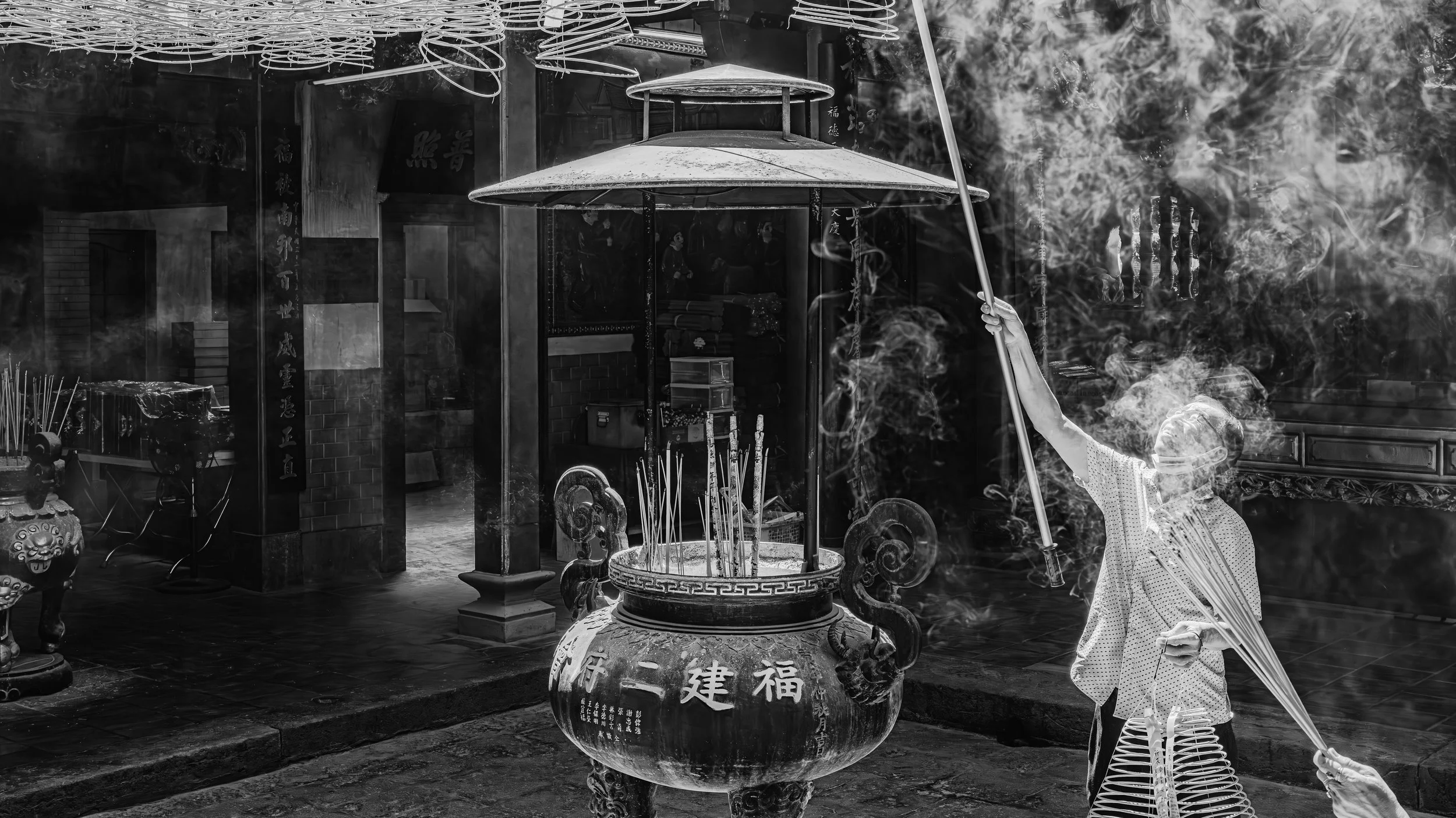 A person burning incense sticks at a shrine with smoke rising, surrounded by traditional Chinese artifacts and decorations.