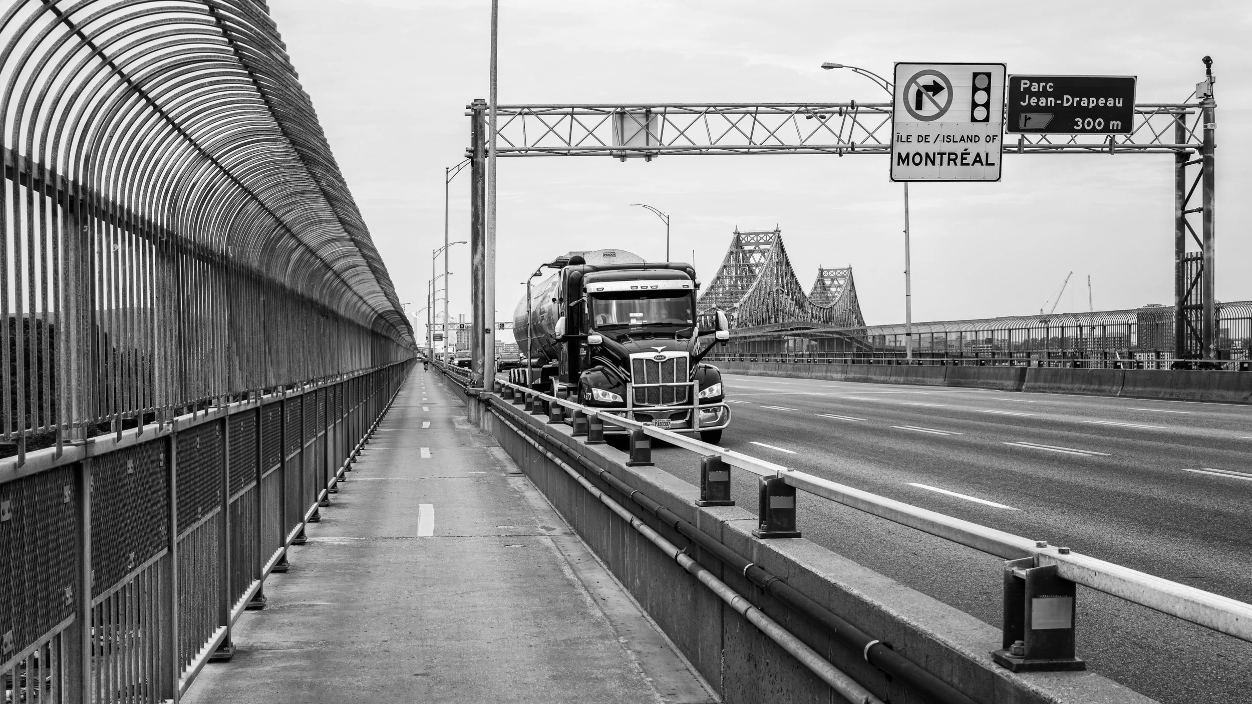 A black-and-white photo of a bridge over a body of water, viewed from a highway with a traffic sign. The bridge has a steel structure with a truss design. A semi-truck is driving on the highway next to a fenced pedestrian walkway. The sign indicates 