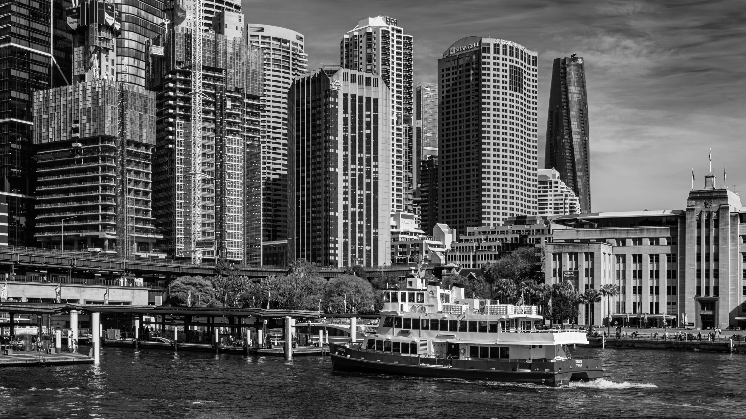 Black and white photo of a city skyline with numerous tall skyscrapers and a ferry boat on the water in the foreground.