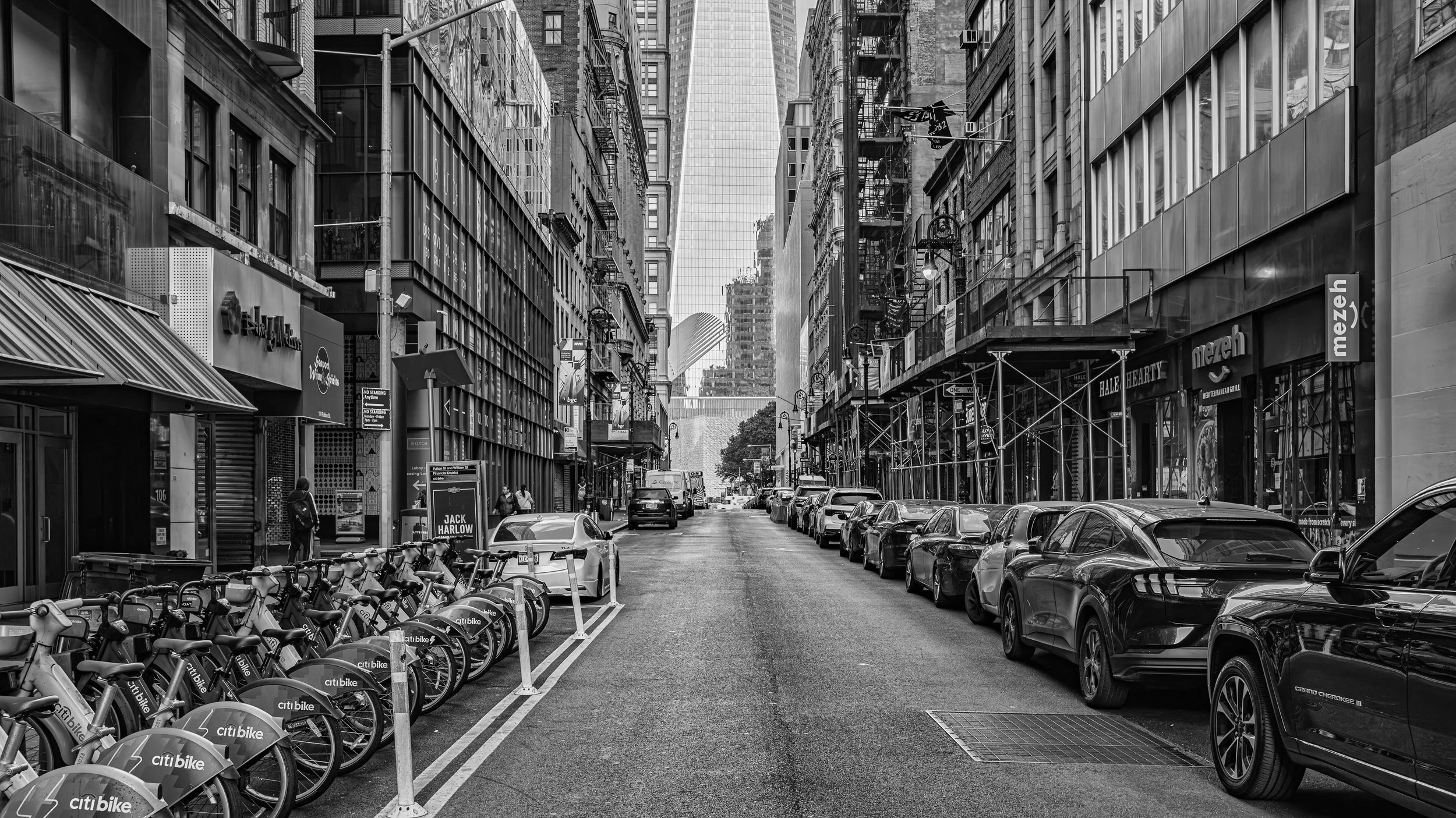 Black and white city street scene with parked cars on the right, bikes on the left, tall buildings, streetlights, and a pedestrian walking. Manhattan, New York, USA.