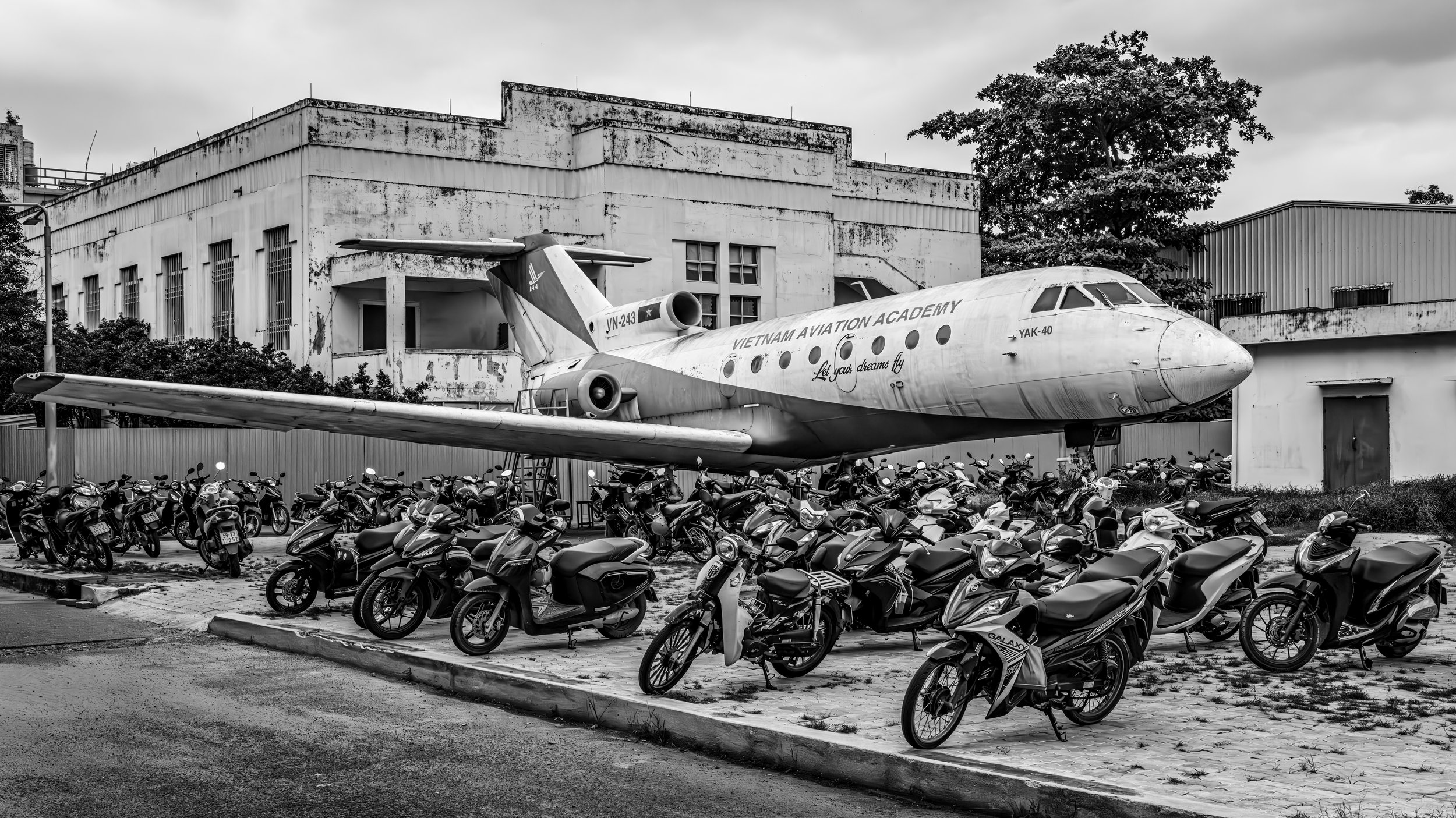 Black and white photo of an airplane on display behind a parking lot filled with motorcycles, with a dilapidated building and trees in the background.