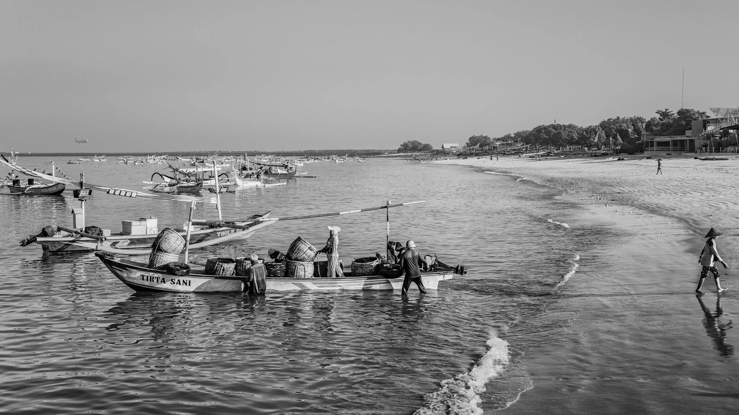 A black and white photo of a coastal scene with boats, fishermen, and buildings on the shoreline.